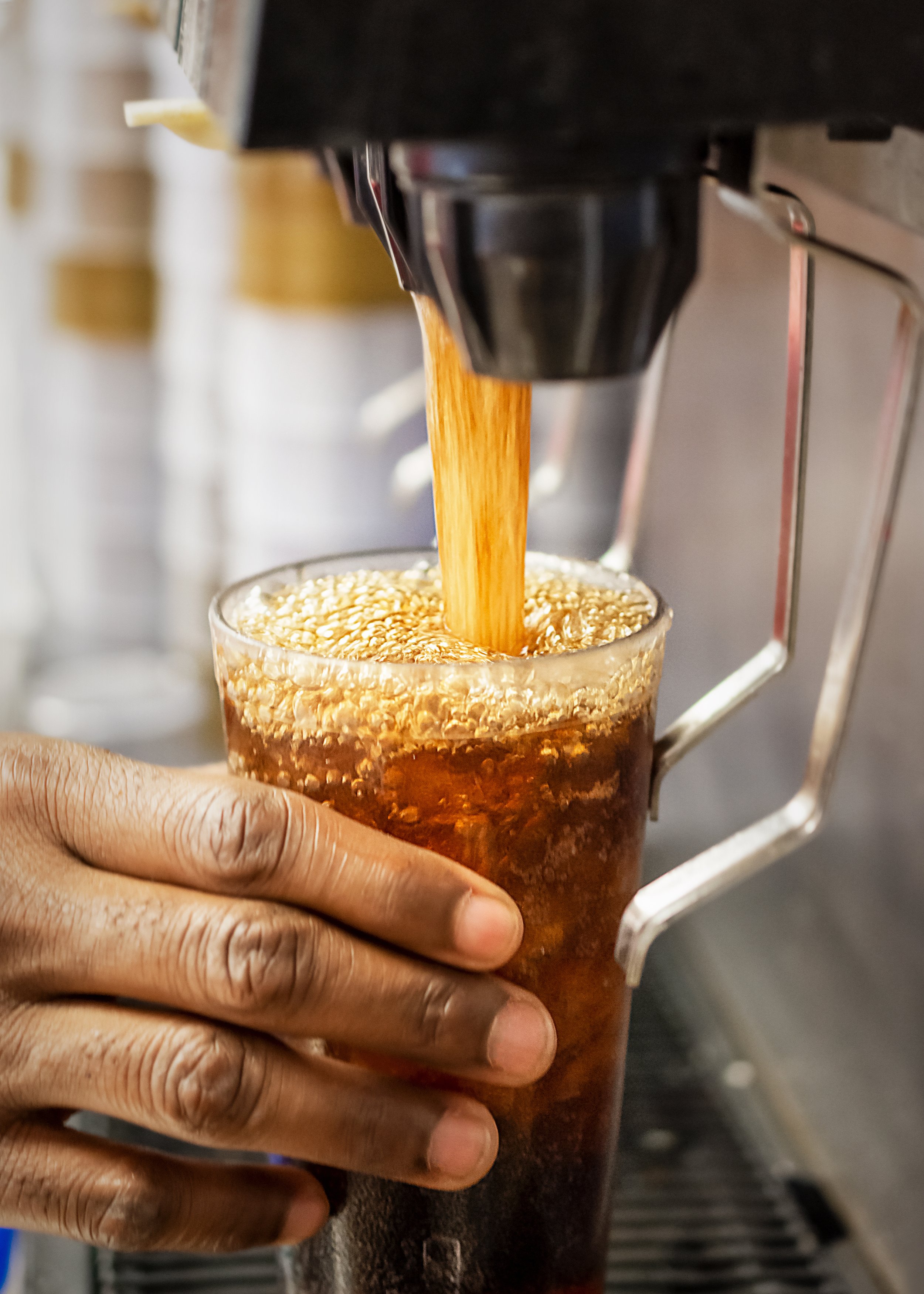 Person's hand holding a glass under a soft drink fountain spout pouring cola into the glass.