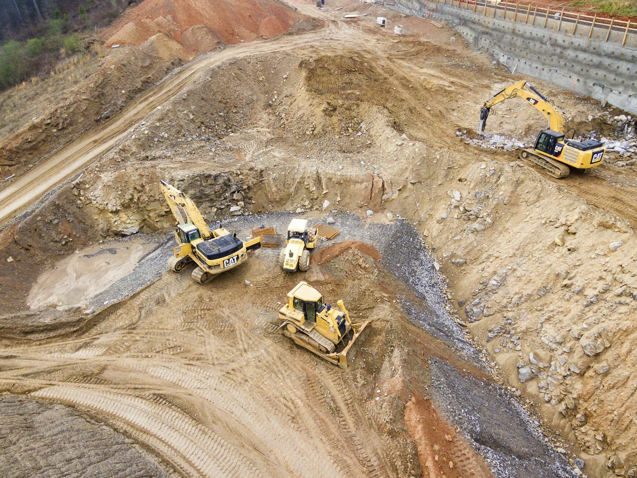 Construction site with excavators digging and moving dirt, with visible tire tracks in the soil and a retaining wall on the side.