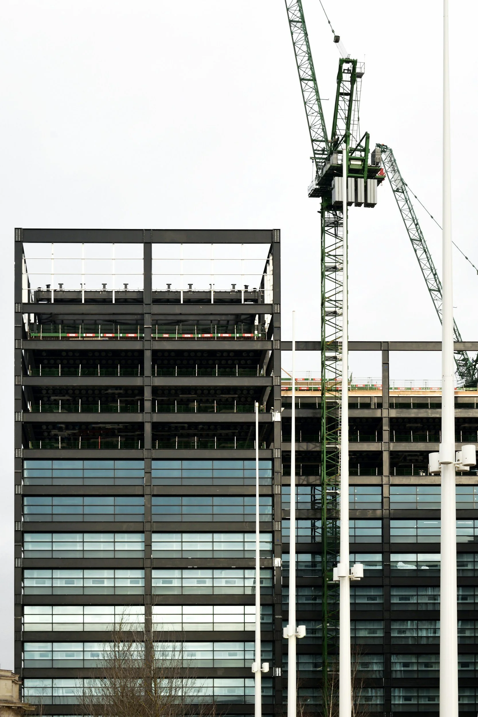 A modern multi-story building under construction with glass windows and metal framework, a construction crane, and flagpoles in front.