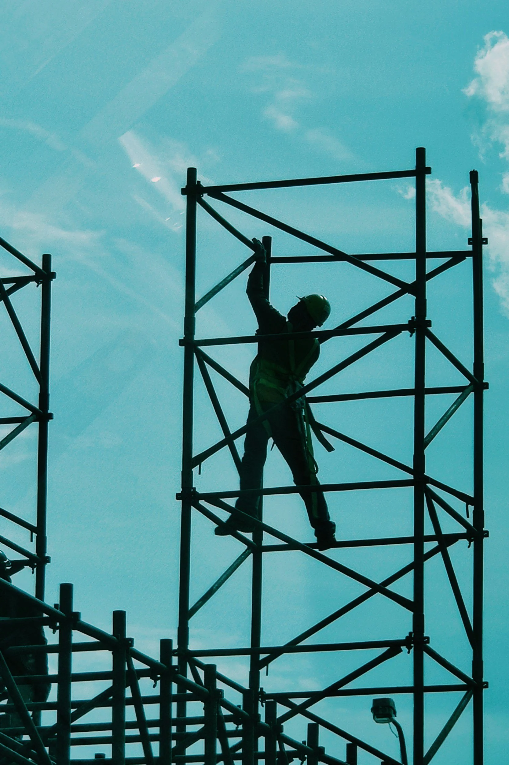 Construction worker wearing a safety helmet climbing scaffolding against a blue sky with clouds.
