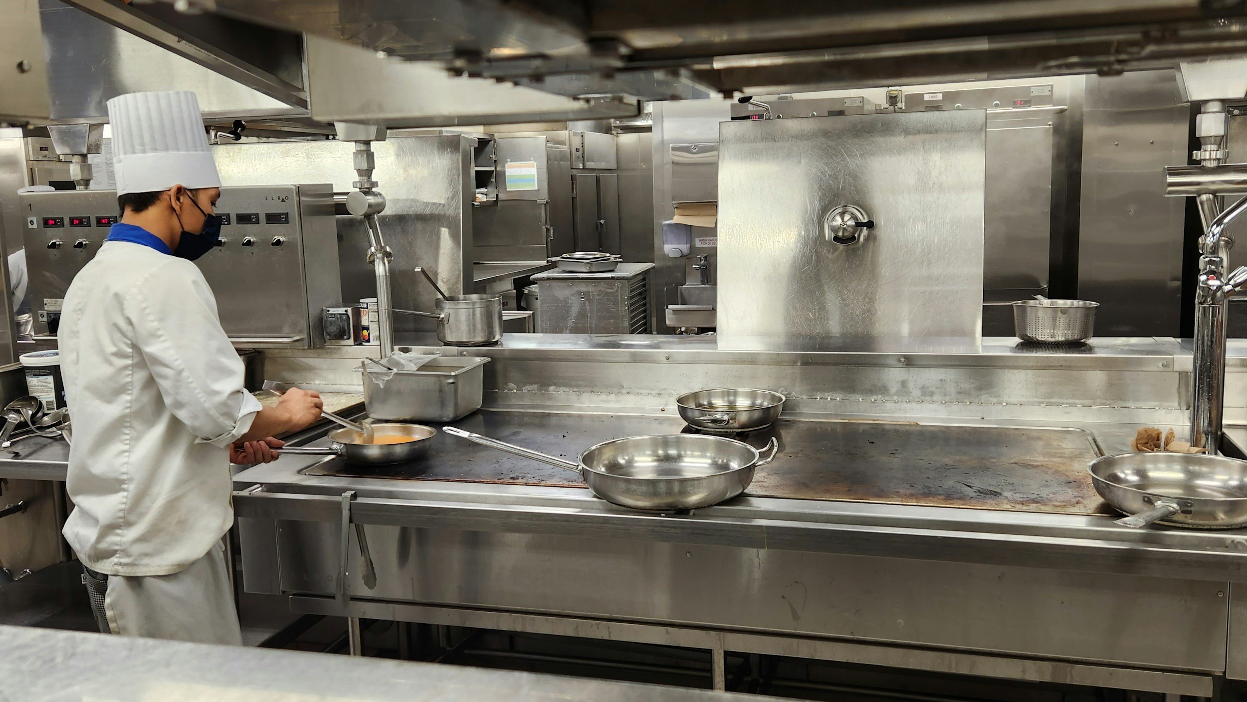 A chef wearing a white uniform, tall hat, and black face mask is cooking in a commercial kitchen. The kitchen has stainless steel appliances and counters, with pots and pans on the stove and prep area.