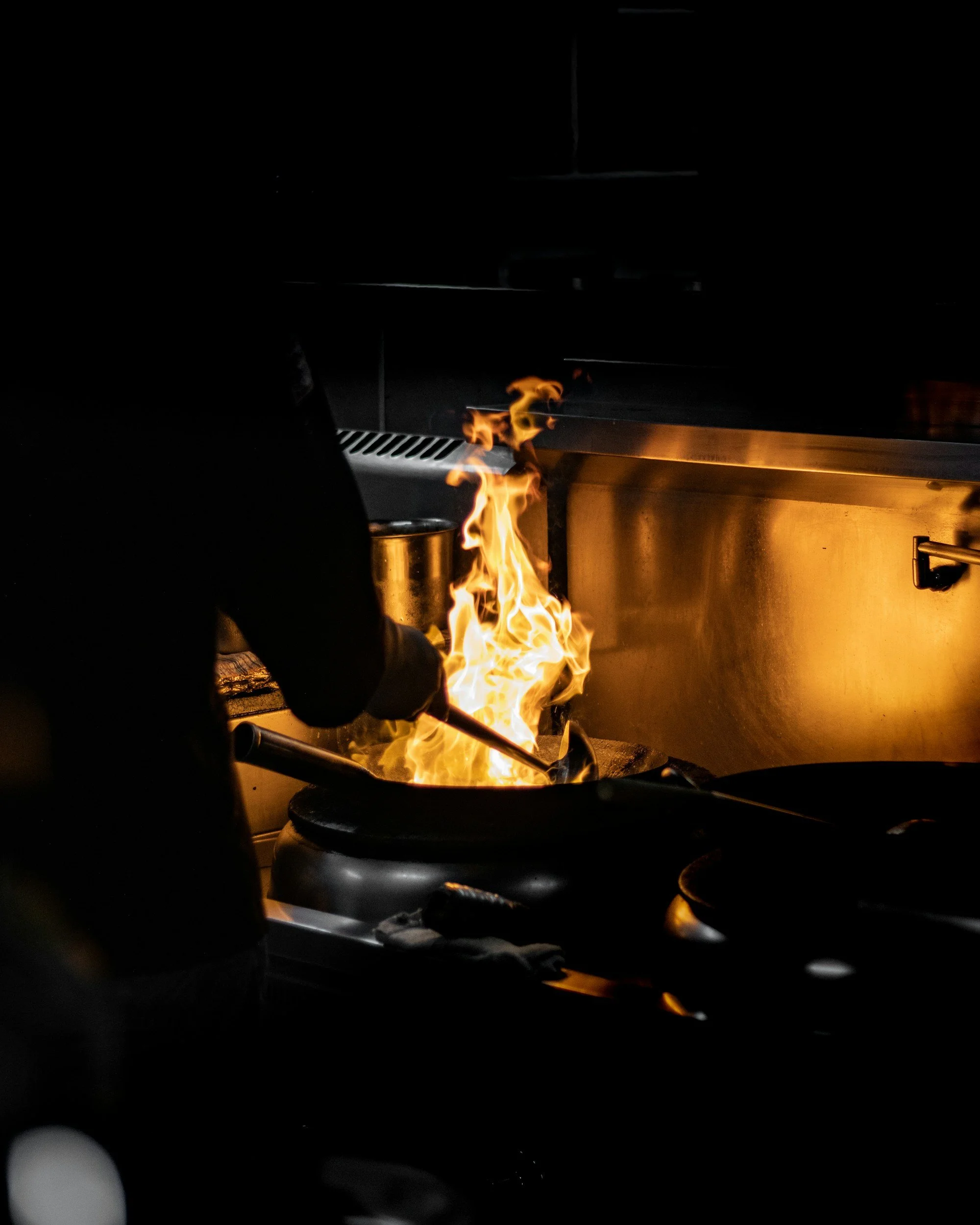 A kitchen stove with a gas flame ignited, producing bright orange and yellow flames, with a visible metallic surface beneath the flames and a dark background.
