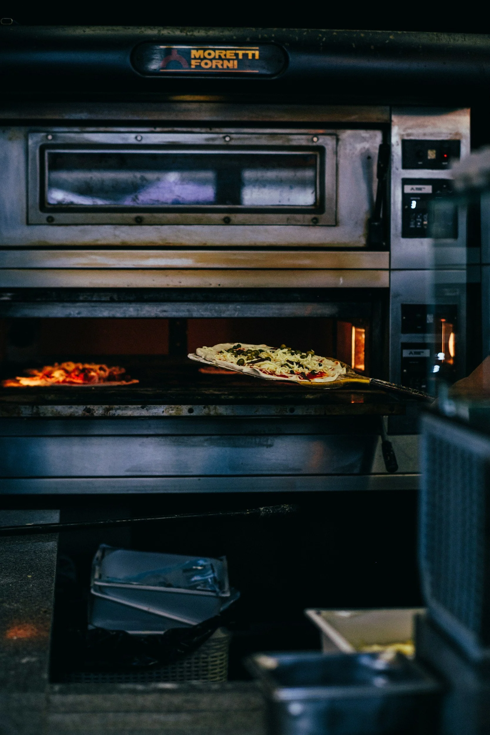 Commercial oven baking a pizza with cheese, black olives, and green peppers, with another pizza partially visible on the left side.