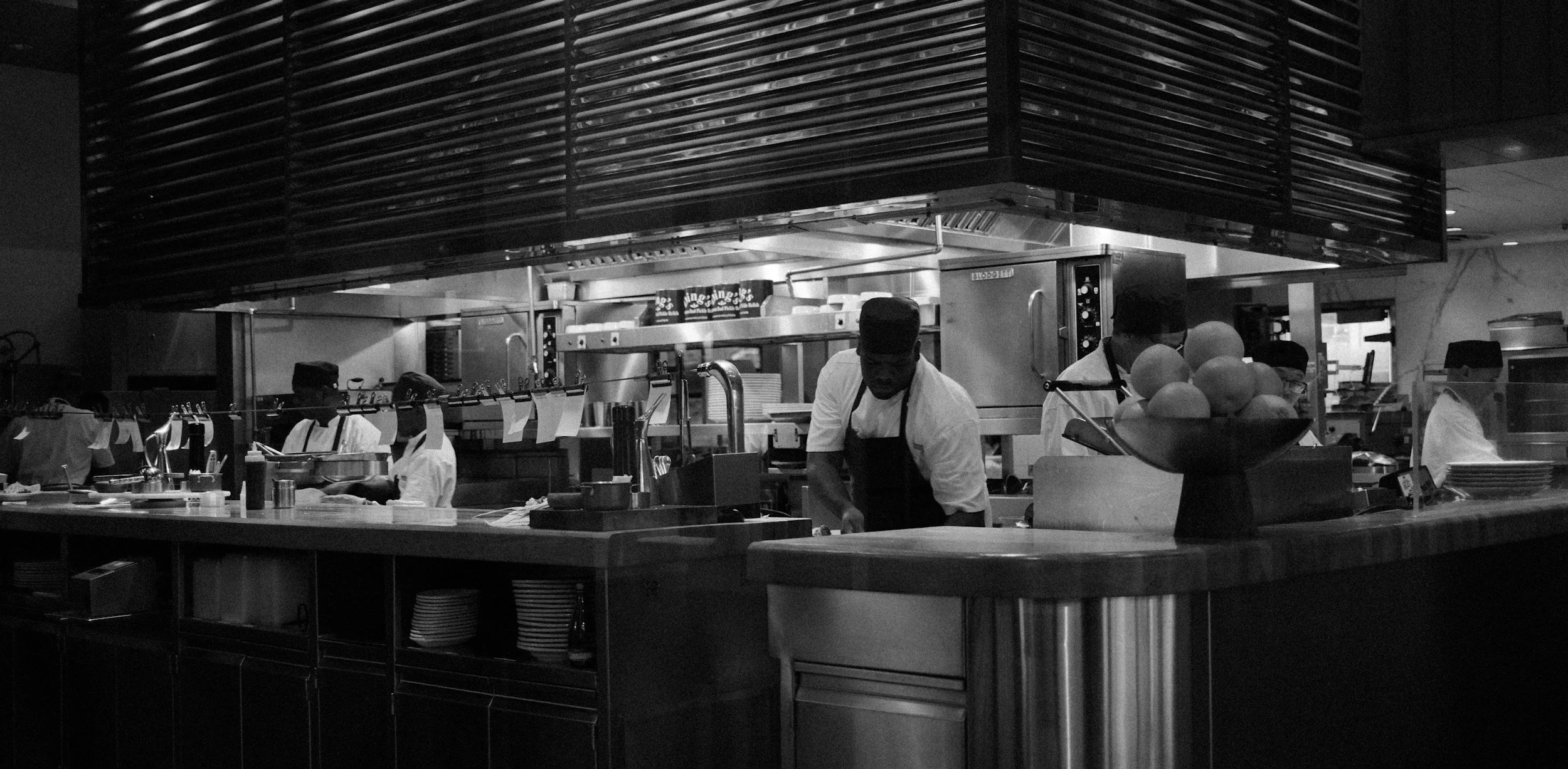 Black and white photo of a commercial kitchen with chefs working behind the counter, stainless steel appliances, and a large bowl of oranges in the foreground.