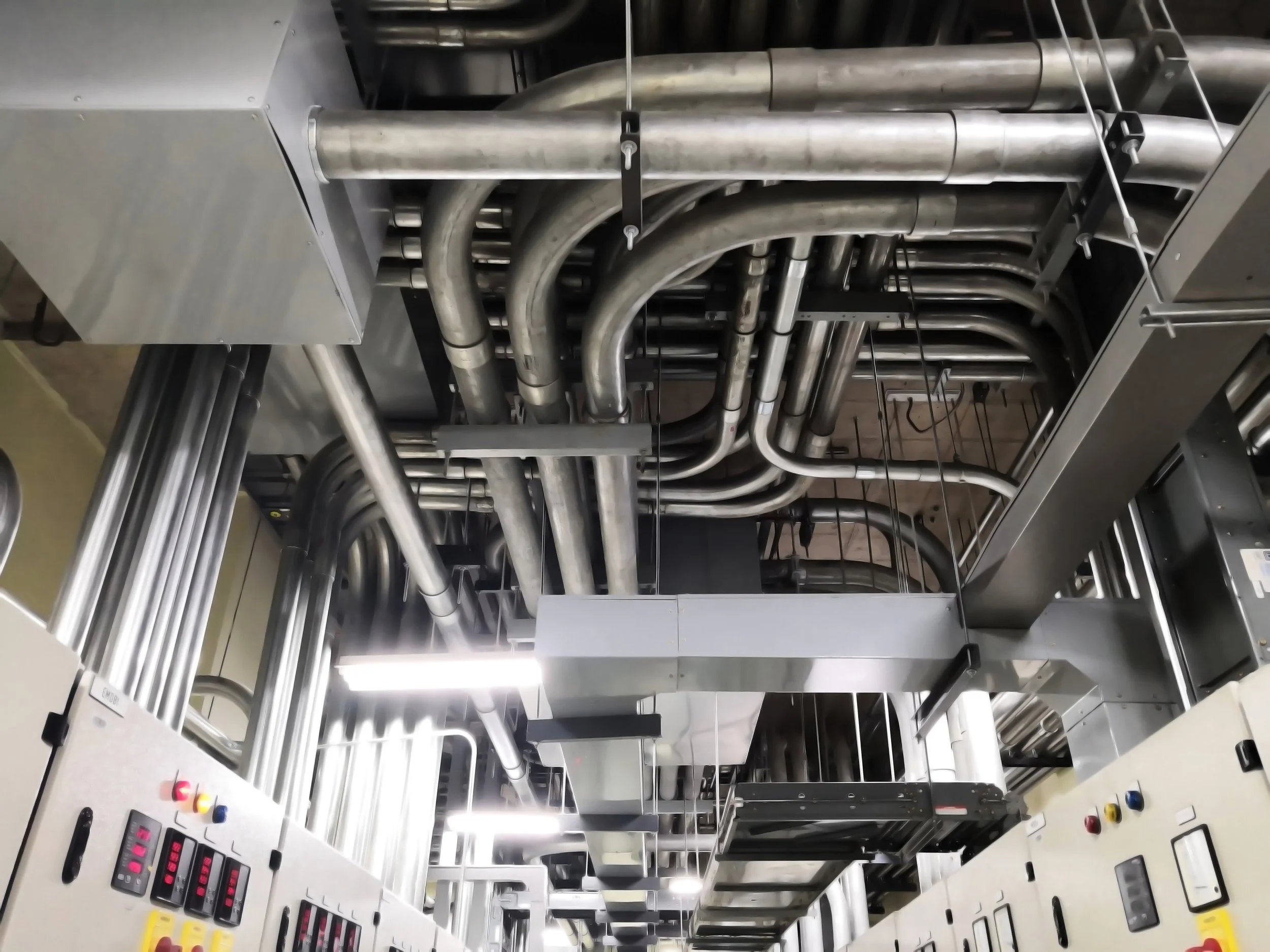 Industrial piping and equipment in a mechanical room or factory setting, with metal pipes, control panels with colored buttons, and structural supports.