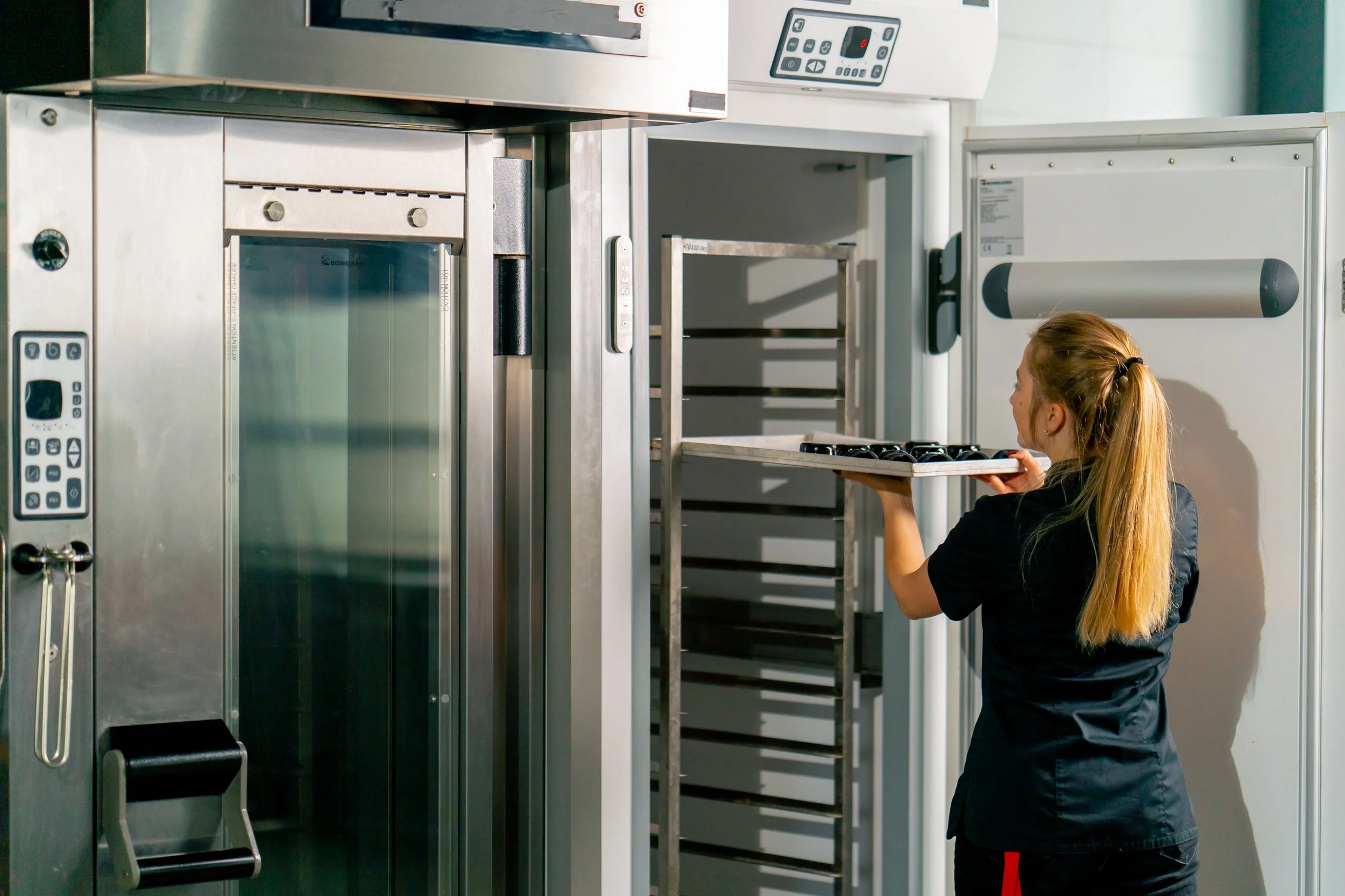 A woman with long blonde hair holding a tray of baked goods in a commercial kitchen or bakery with stainless steel appliances and shelving.