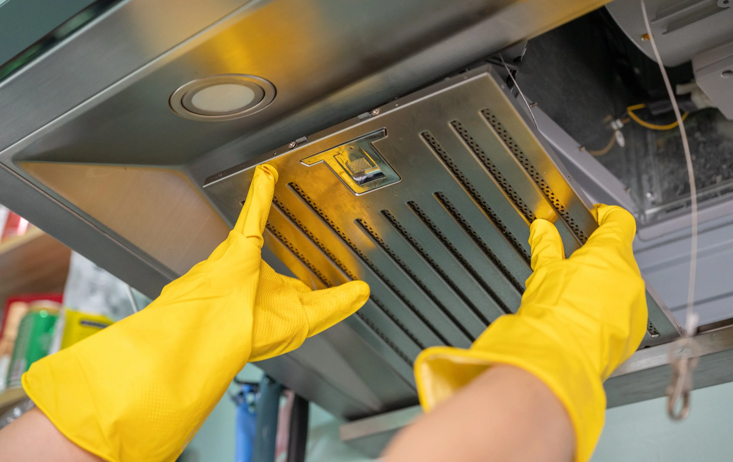 Hands in yellow gloves working on metallic kitchen appliance or industrial equipment under a metal surface.