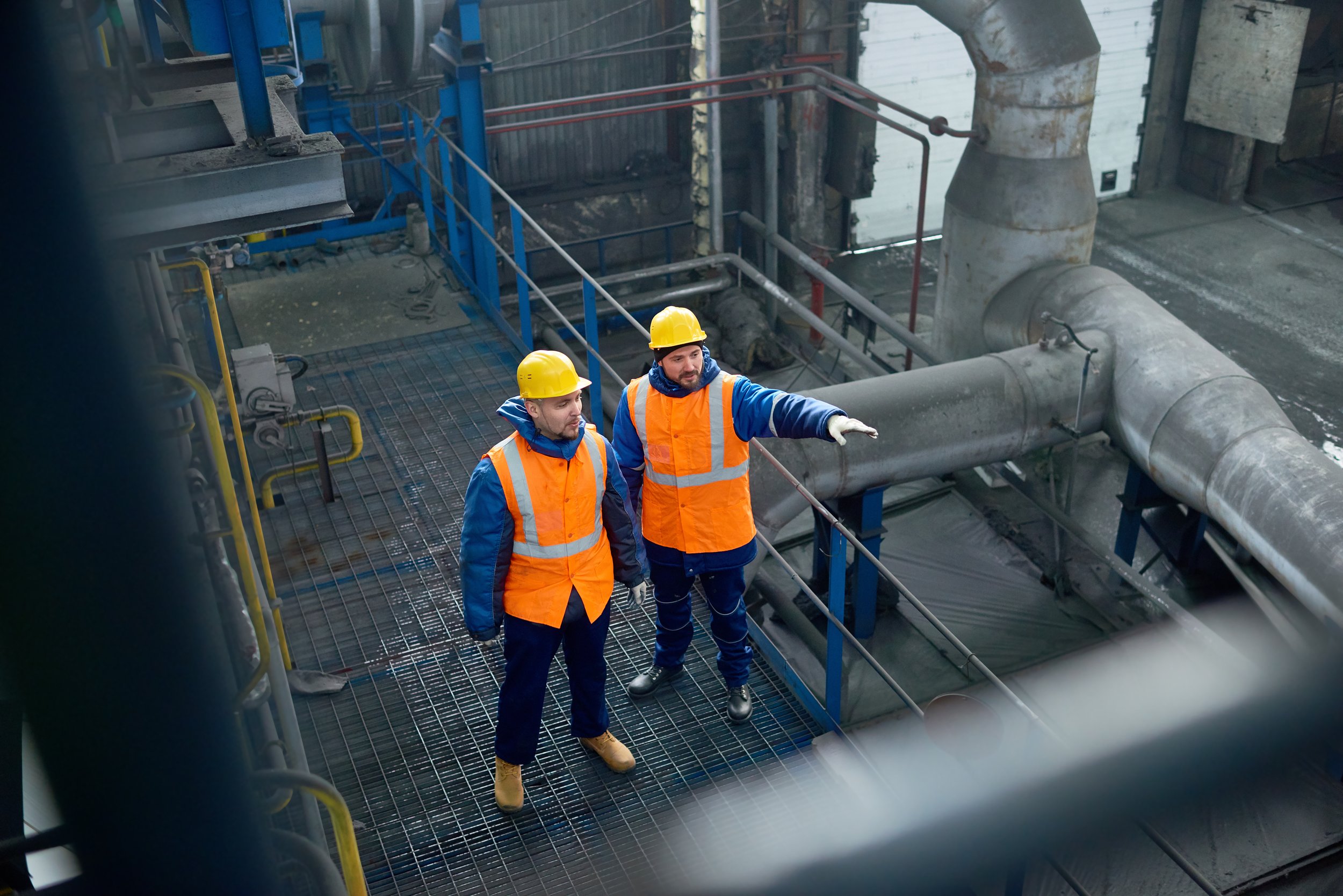Two male workers wearing yellow safety helmets, orange safety vests, and gloves are standing on a metal grate platform and inspecting large industrial pipes in an industrial facility.