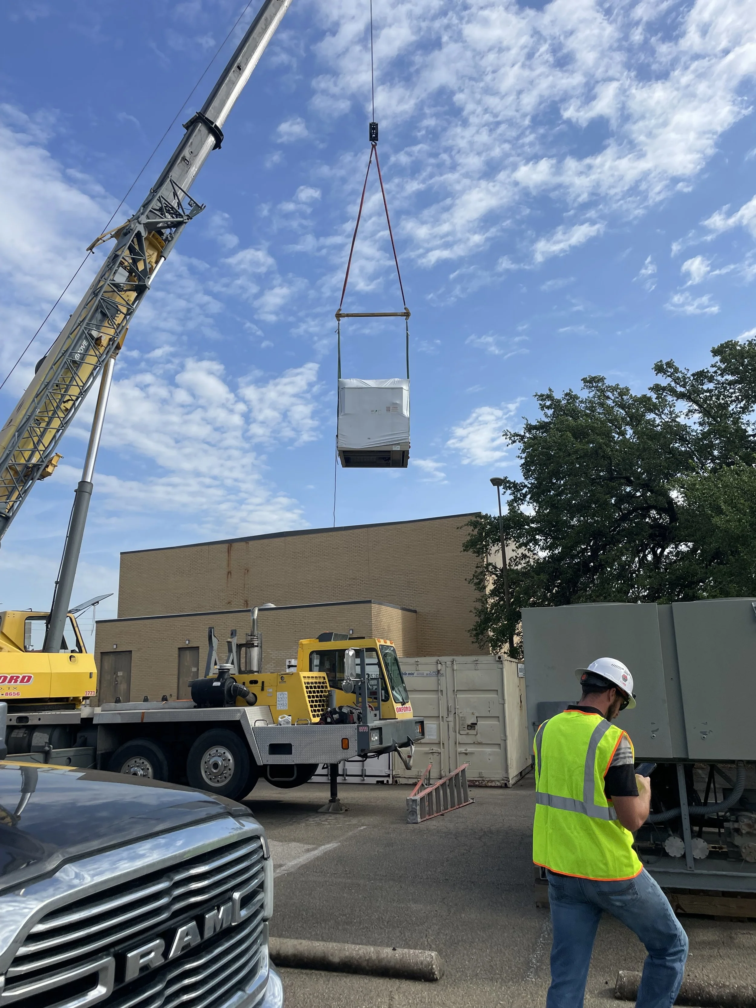 A construction site with a large crane lifting an air conditioning unit, workers in safety vests and helmets, parked vehicles, and a brick building in the background under a partly cloudy sky.
