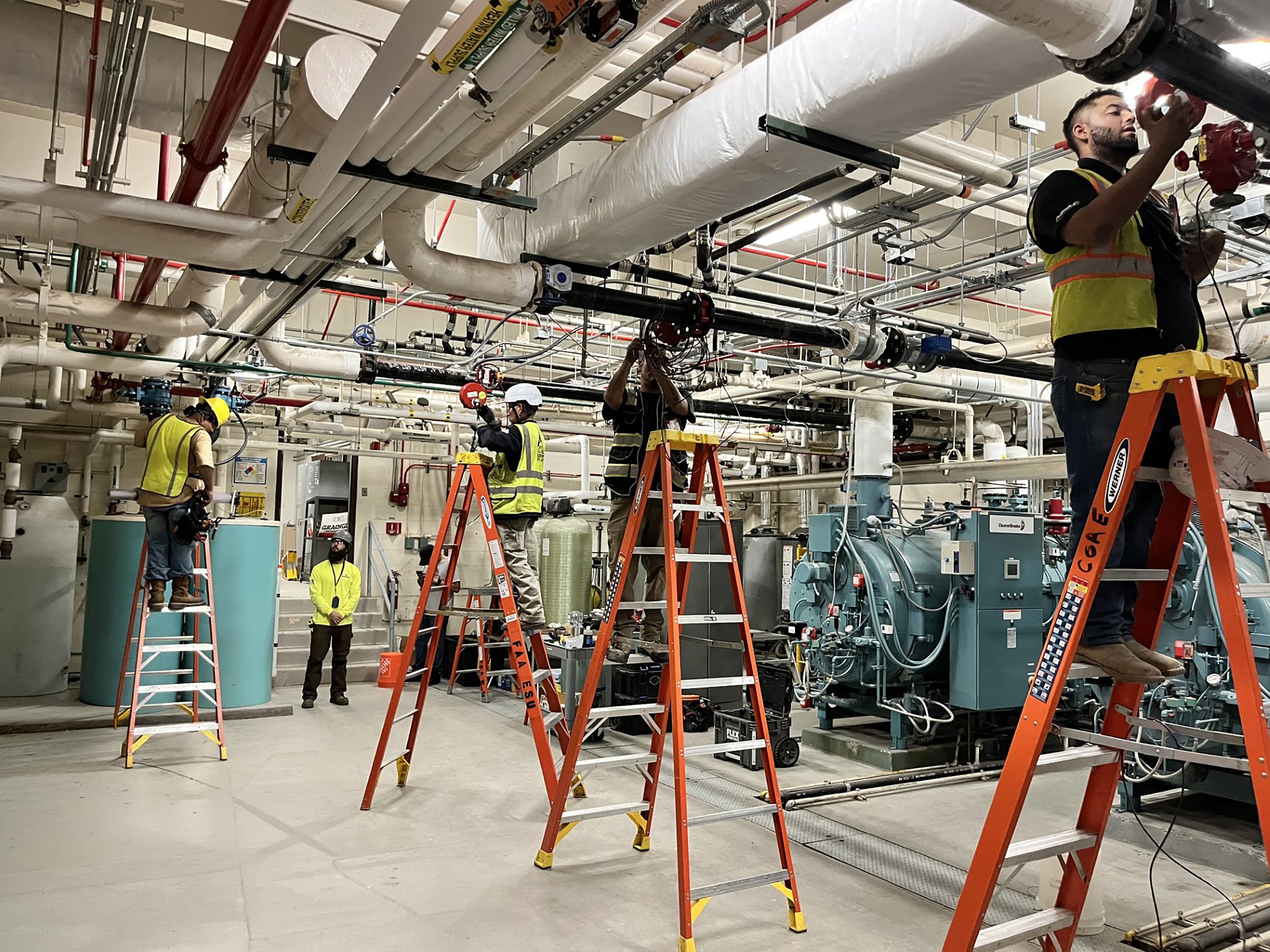 Industrial setting with workers on ladders inspecting and working on large pipes and machinery, wearing safety gear including helmets and vests.