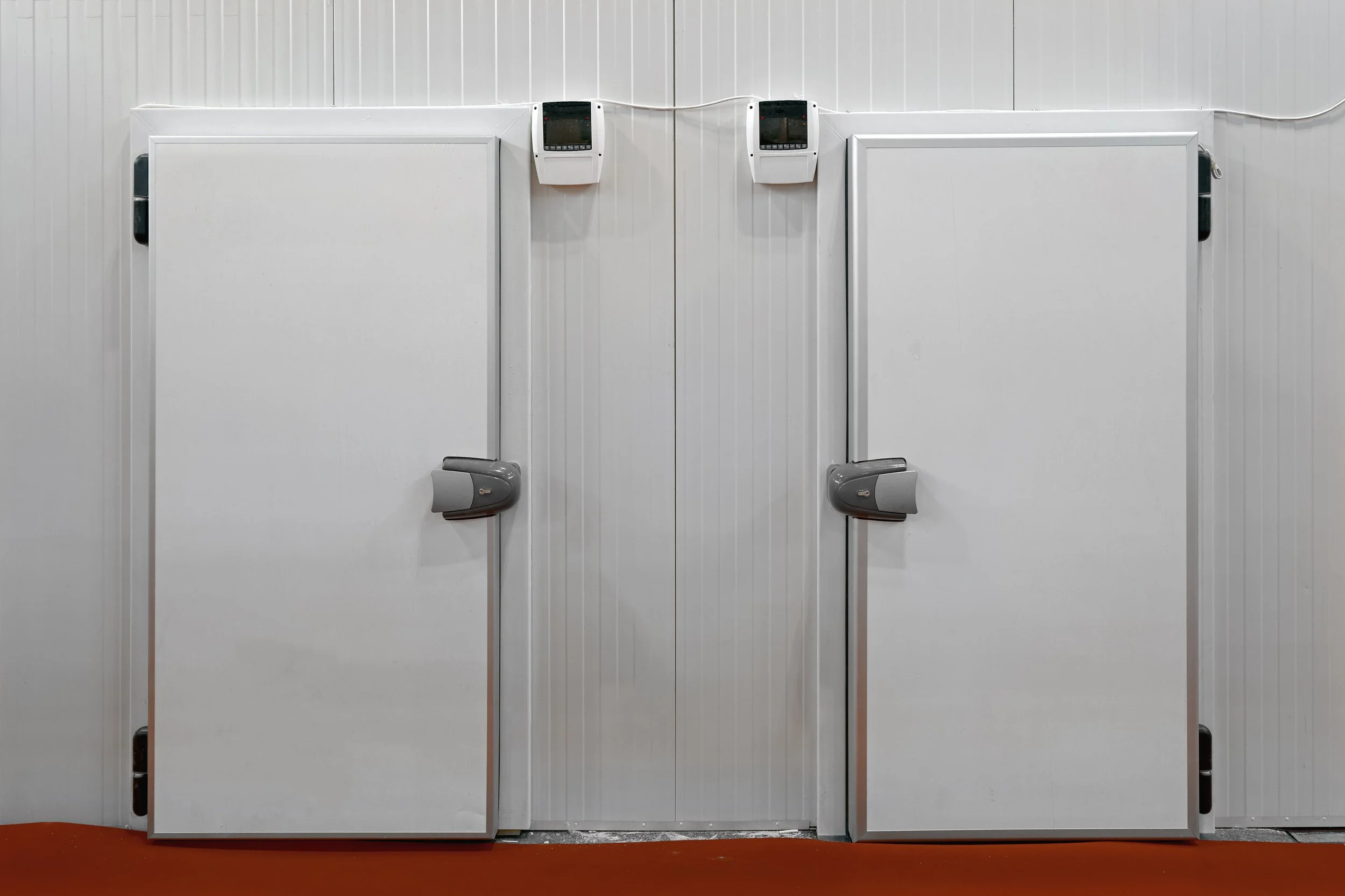 Two white bathroom stalls with closed doors, each with a gray automatic hand dryer mounted on the wall above. The stalls are separated by a narrow gap and are mounted to a red floor with a white wall background.