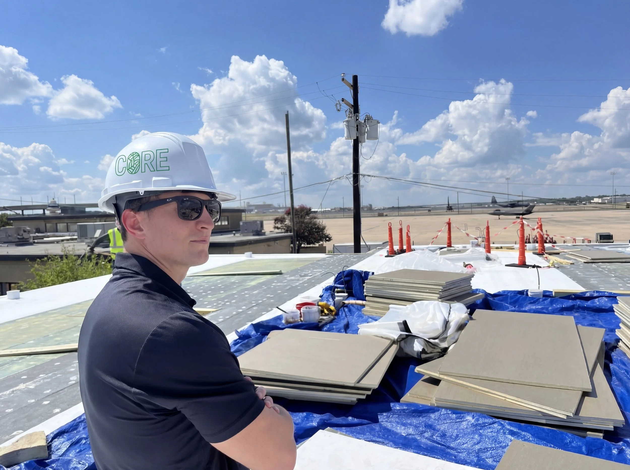 A man in a white hard hat with the word 'CORE' and a logo on it, and sunglasses, is sitting outdoors at a construction or industrial site under a partly cloudy sky, with a large aircraft in the background and construction materials and equipment around him.