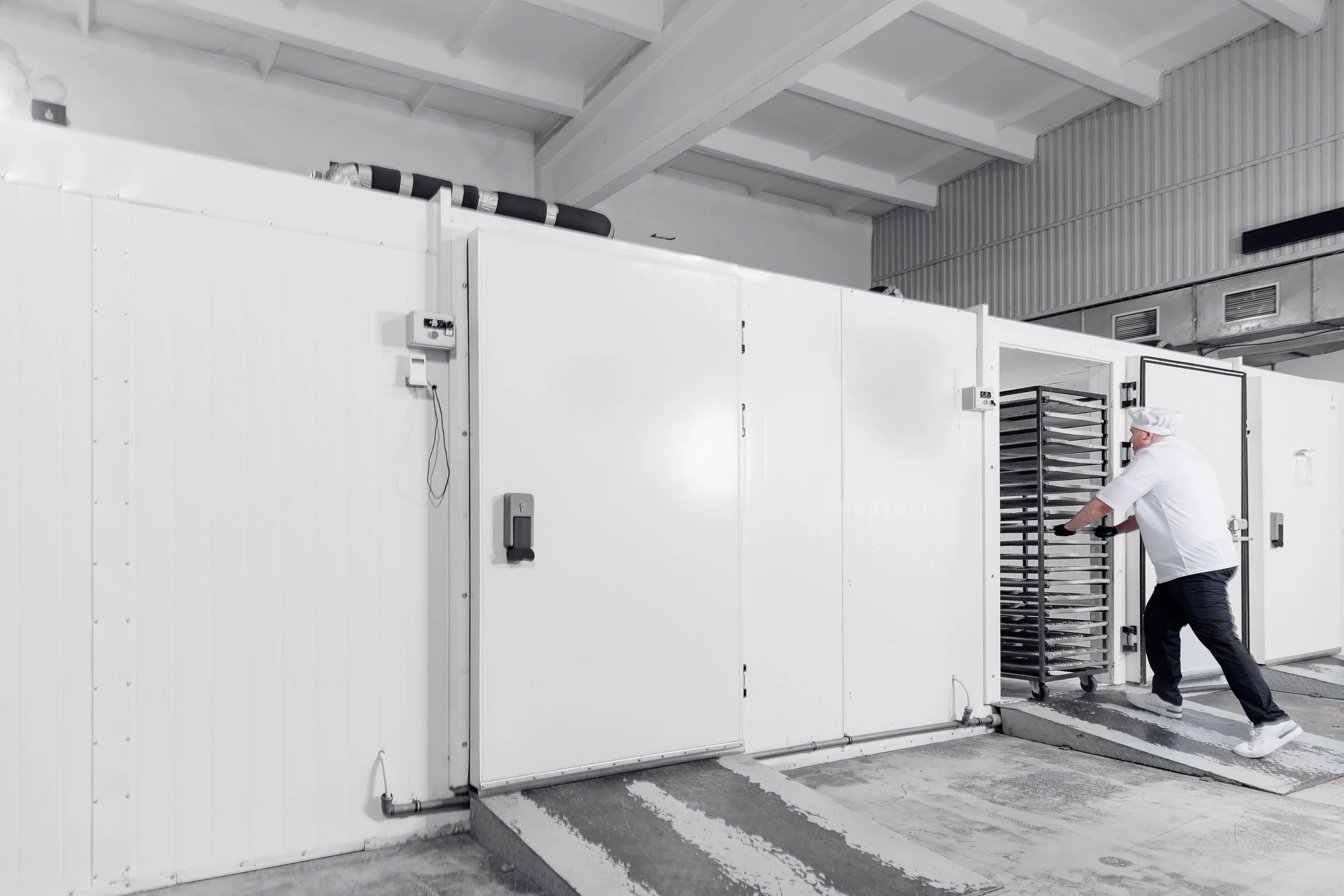 A person dressed as a chef, wearing a white chef's coat and hat, standing in front of a large industrial refrigeration unit, working with a tray rack inside the unit.