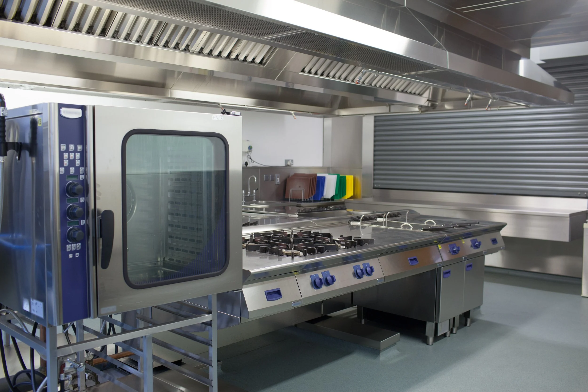 Commercial kitchen with stainless steel appliances, including an oven, stovetop, ventilation hood, and colorful cutting boards.