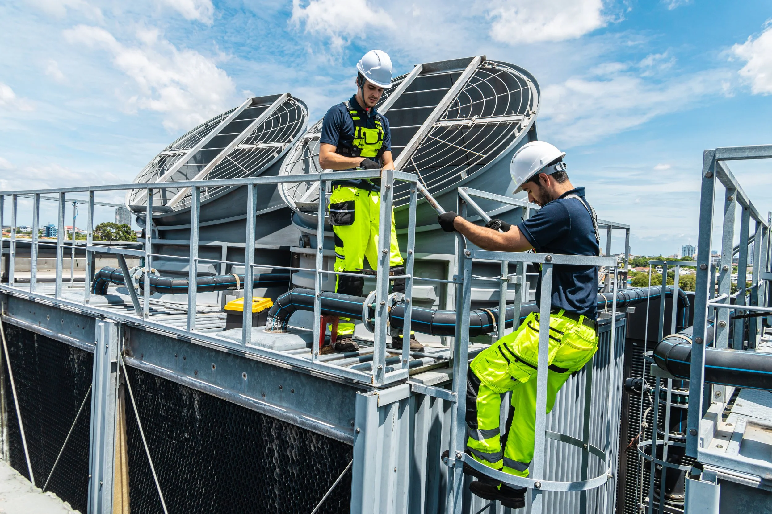 Two male workers in safety helmets and reflective clothing working on HVAC equipment on a rooftop.
