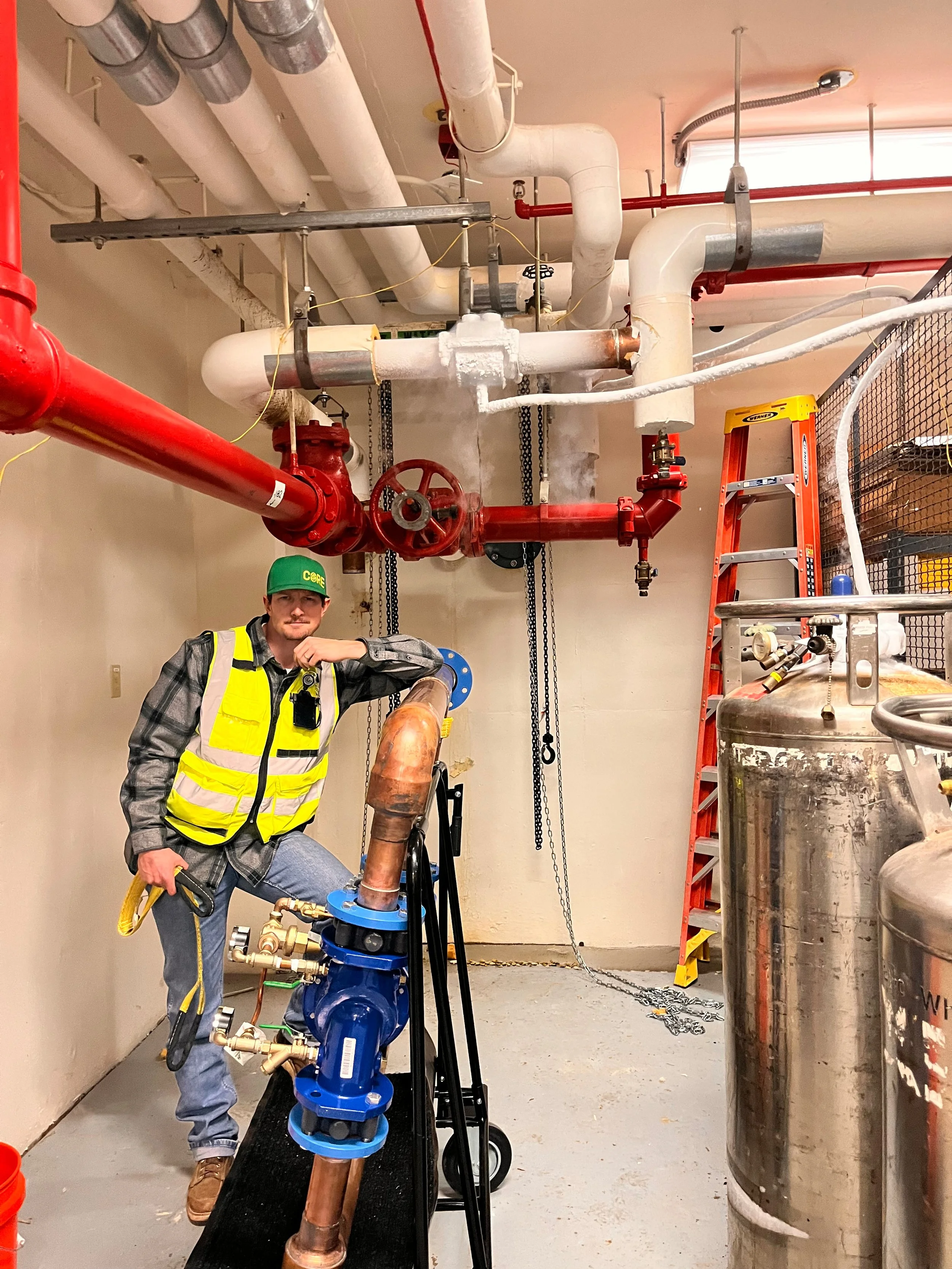 A man in a yellow safety vest and green hard hat posing next to plumbing equipment in an industrial setting, with large pipes, valves, and a ladder in the background.