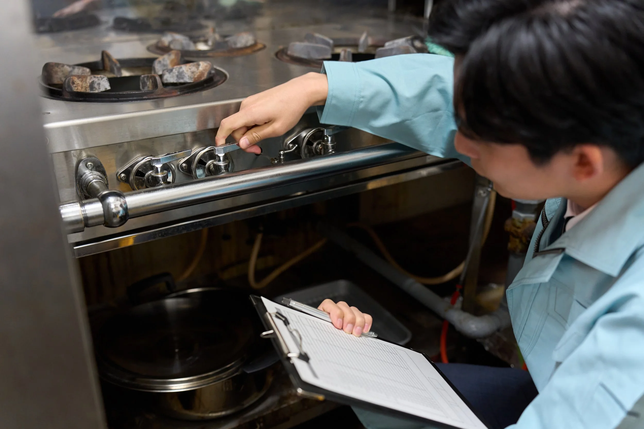 Person in blue coat adjusting stove knobs in commercial kitchen while taking notes on clipboard.