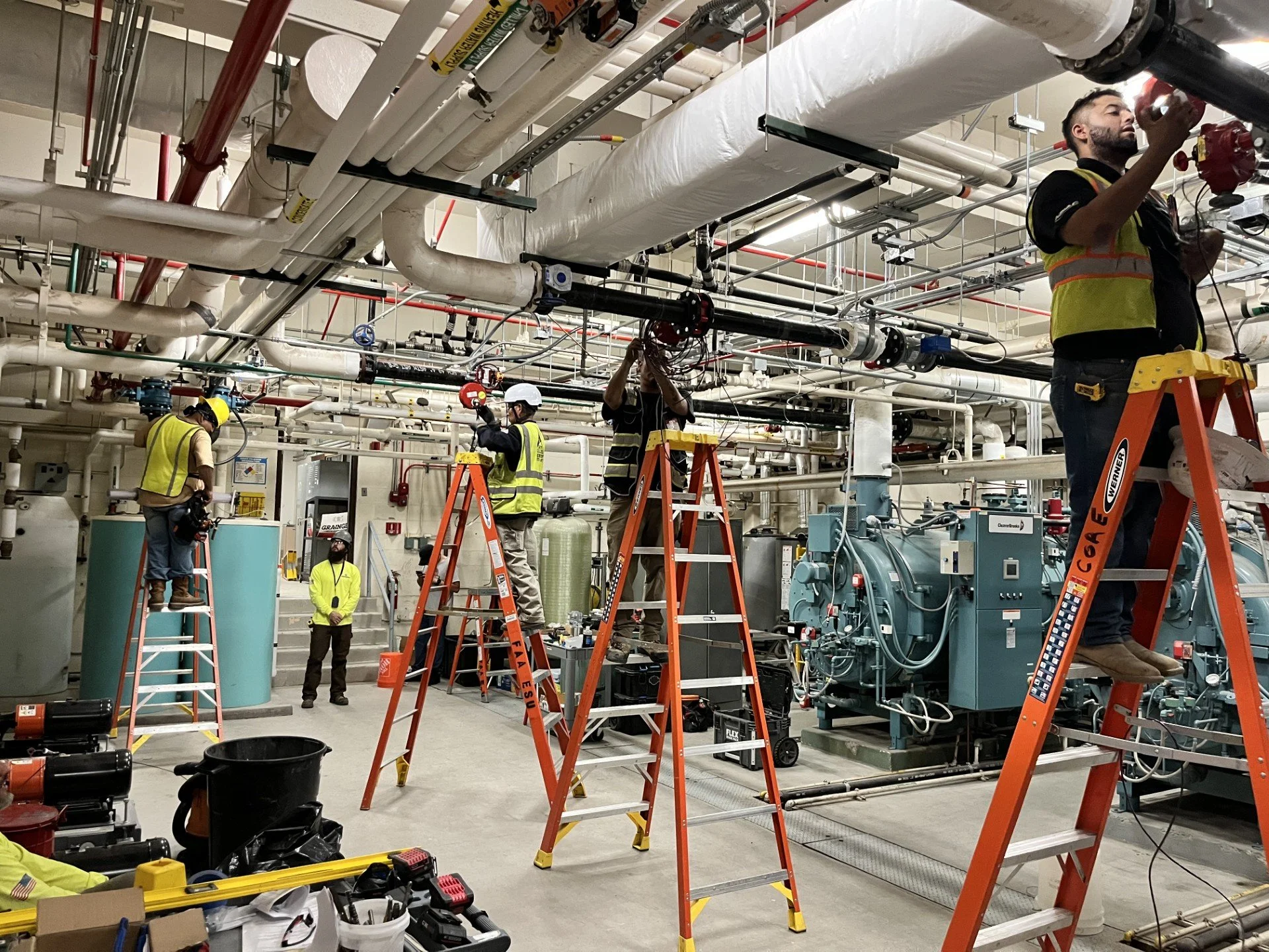 Workers in safety vests and helmets working on industrial pipes and machinery, using ladders in a mechanical room