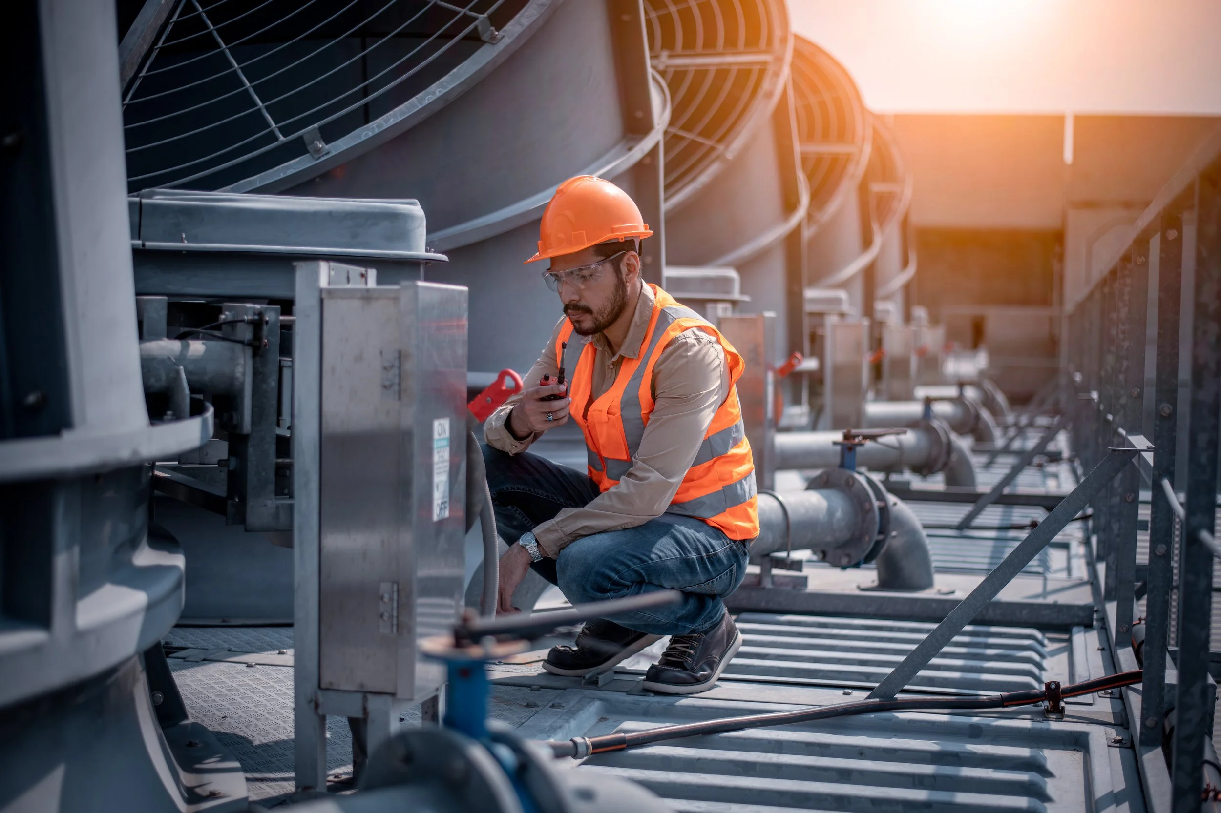 A male worker wearing safety glasses, an orange hard hat, and a reflective vest inspecting industrial equipment outdoors during sunset.