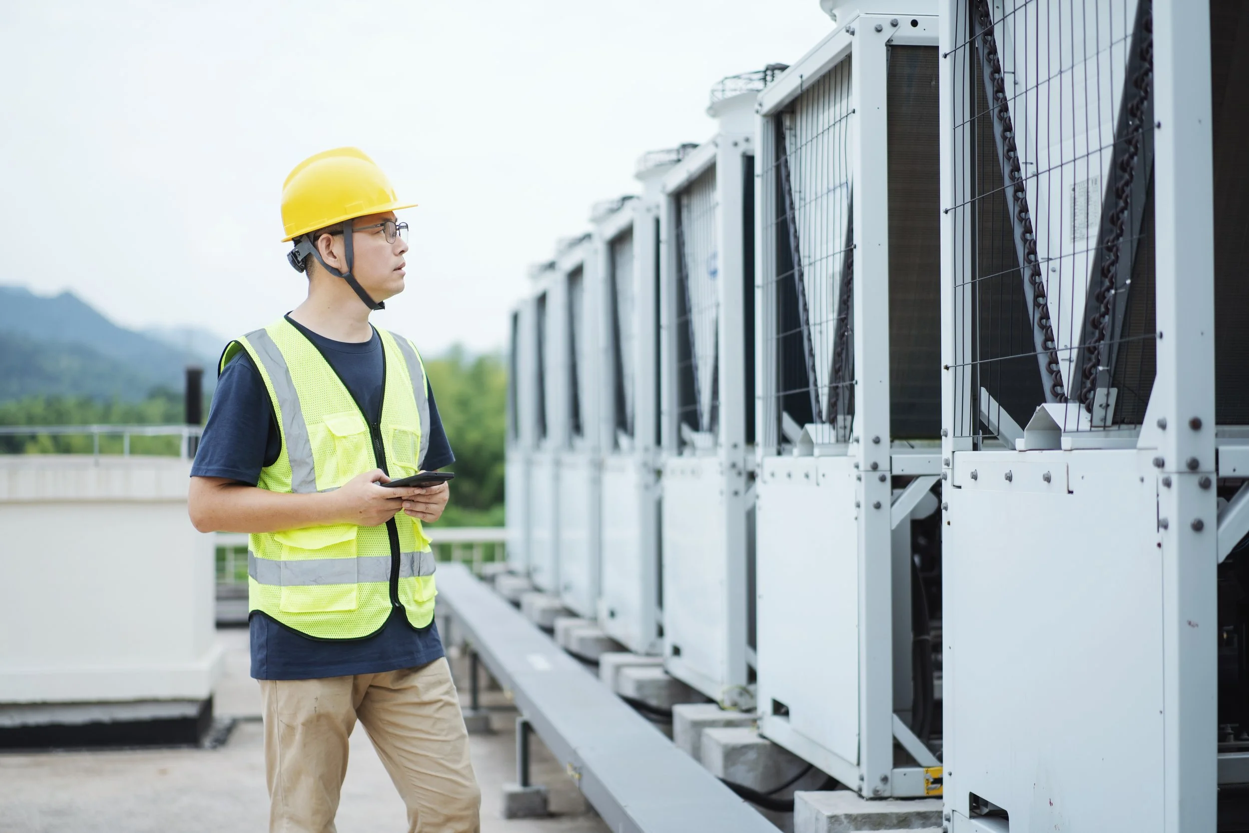 Man wearing a yellow safety helmet and reflective vest inspecting industrial equipment on a rooftop with a mountainous background.