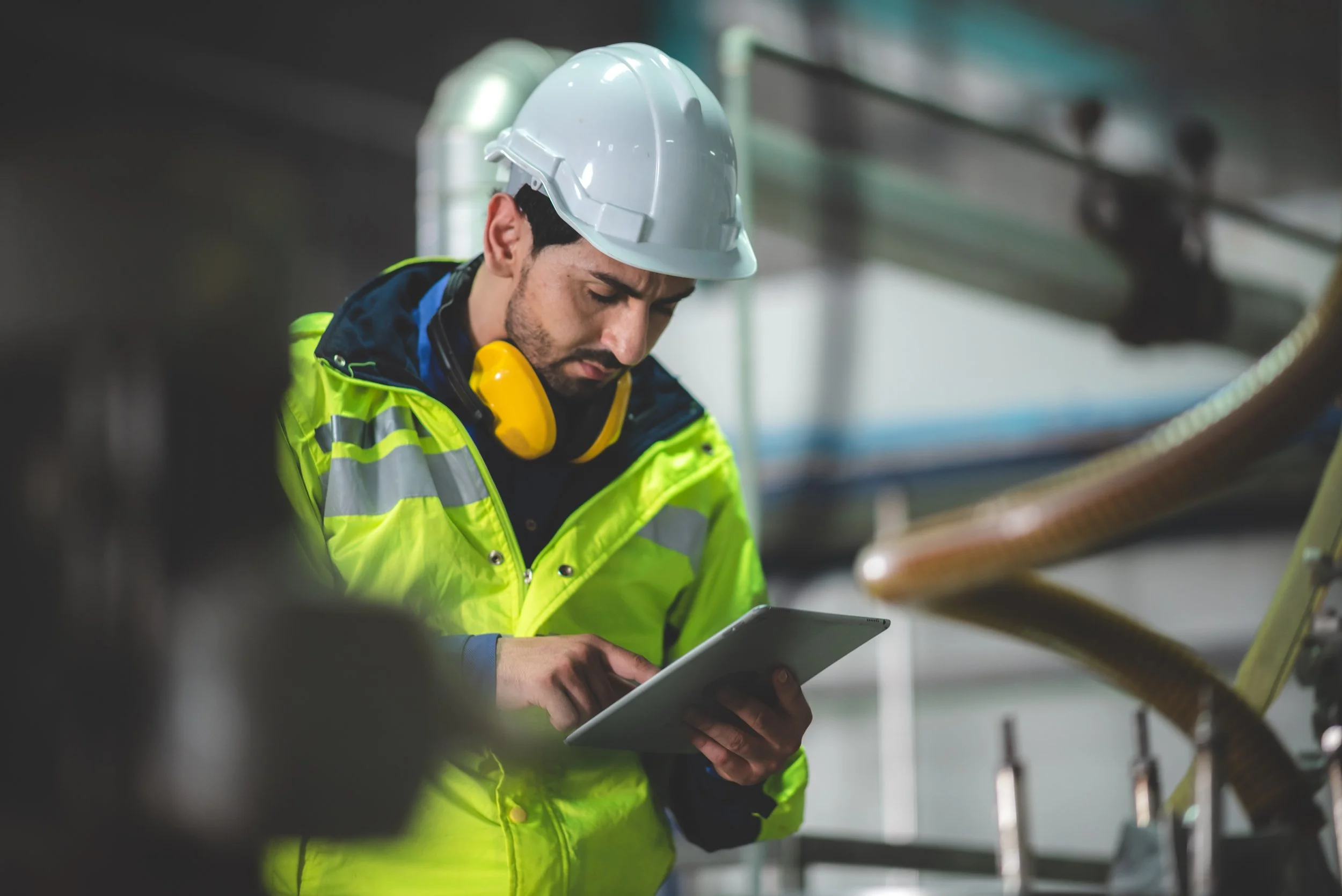 A male industrial worker wearing a white helmet, yellow ear protection, a high-visibility green jacket with reflective stripes, and dark clothing, working and inspecting equipment with a tablet device in a factory or industrial setting.