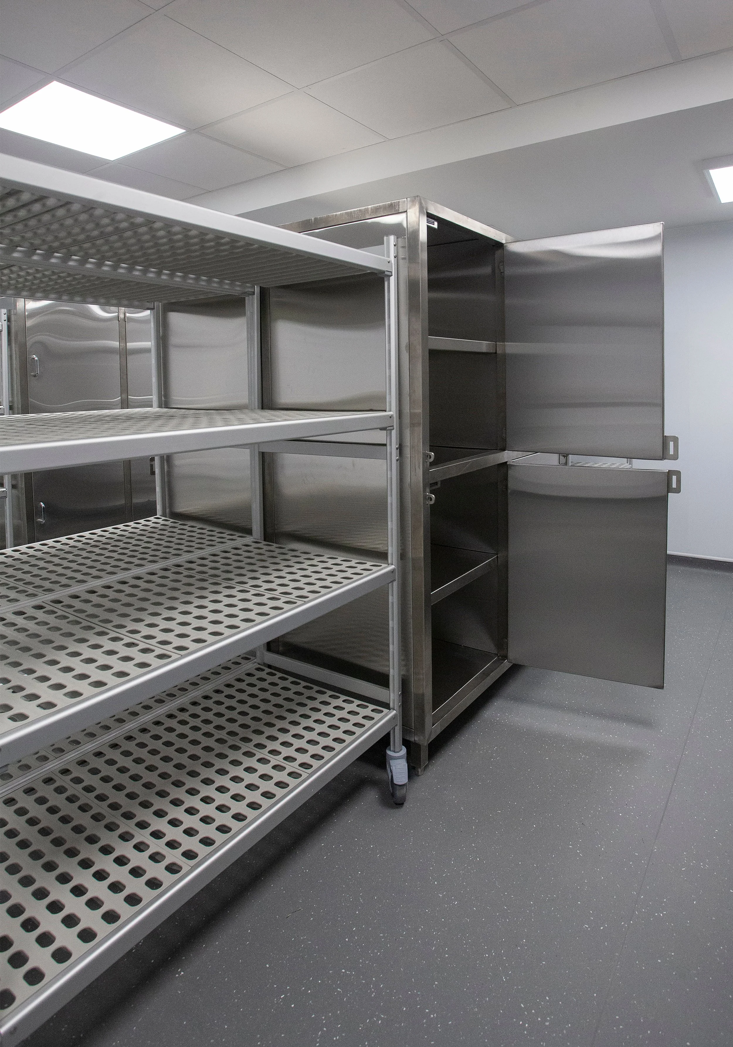 Empty stainless steel shelving unit and storage cabinets in a clean, well-lit industrial or laboratory room with grey speckled floor and ceiling lights.