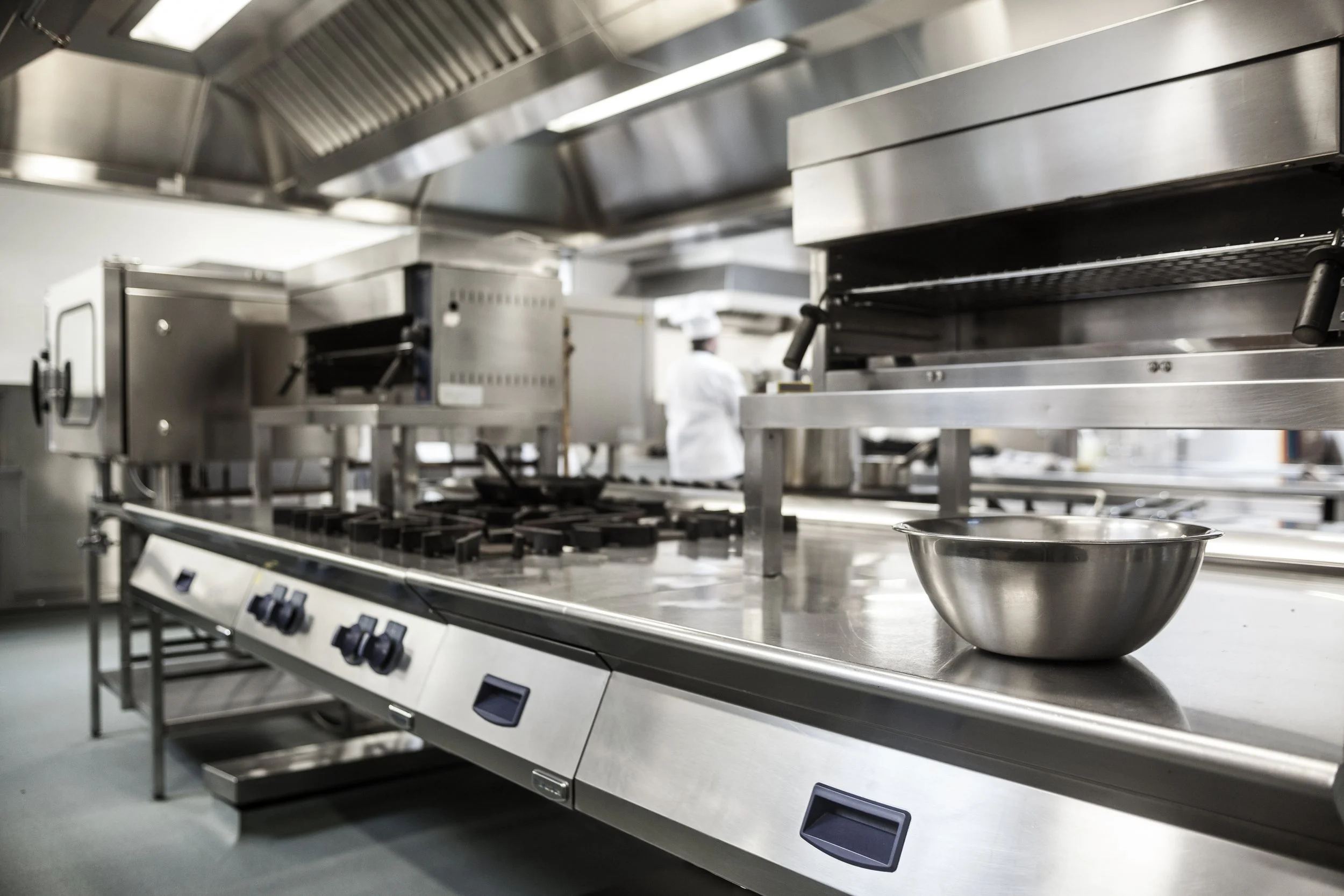 Commercial kitchen with stainless steel stoves and a metal mixing bowl in the foreground.