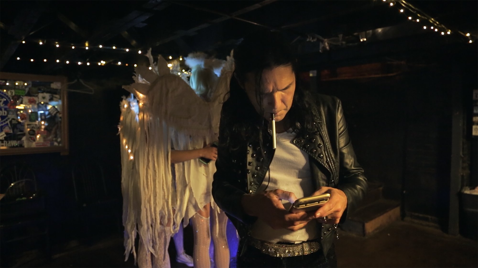 A man with long black hair wearing a leather jacket and white t-shirt, looking at his phone, with a cigarette in his mouth, standing in a dimly lit room with string lights overhead, and a person dressed as an angel in the background.