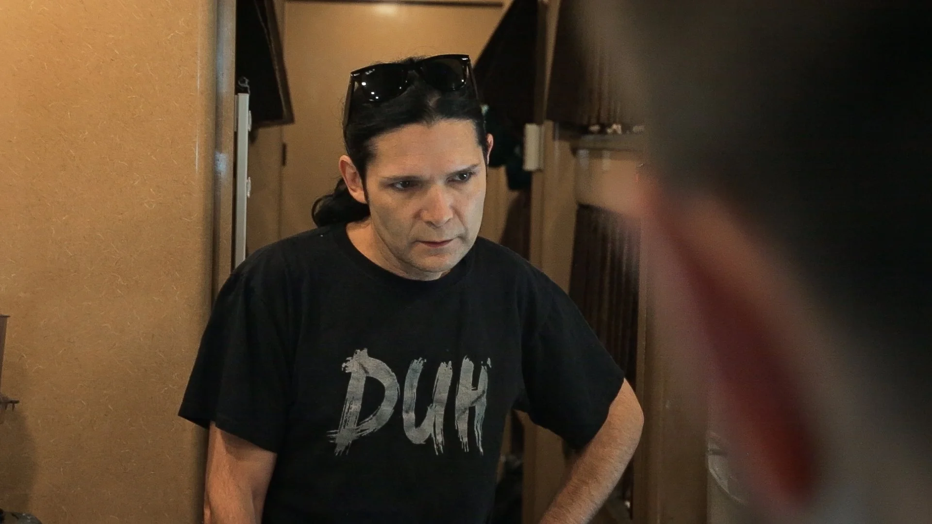 Corey Feldman is wearing a black T-shirt with the word 'DUH' on it, standing indoors with a serious expression.