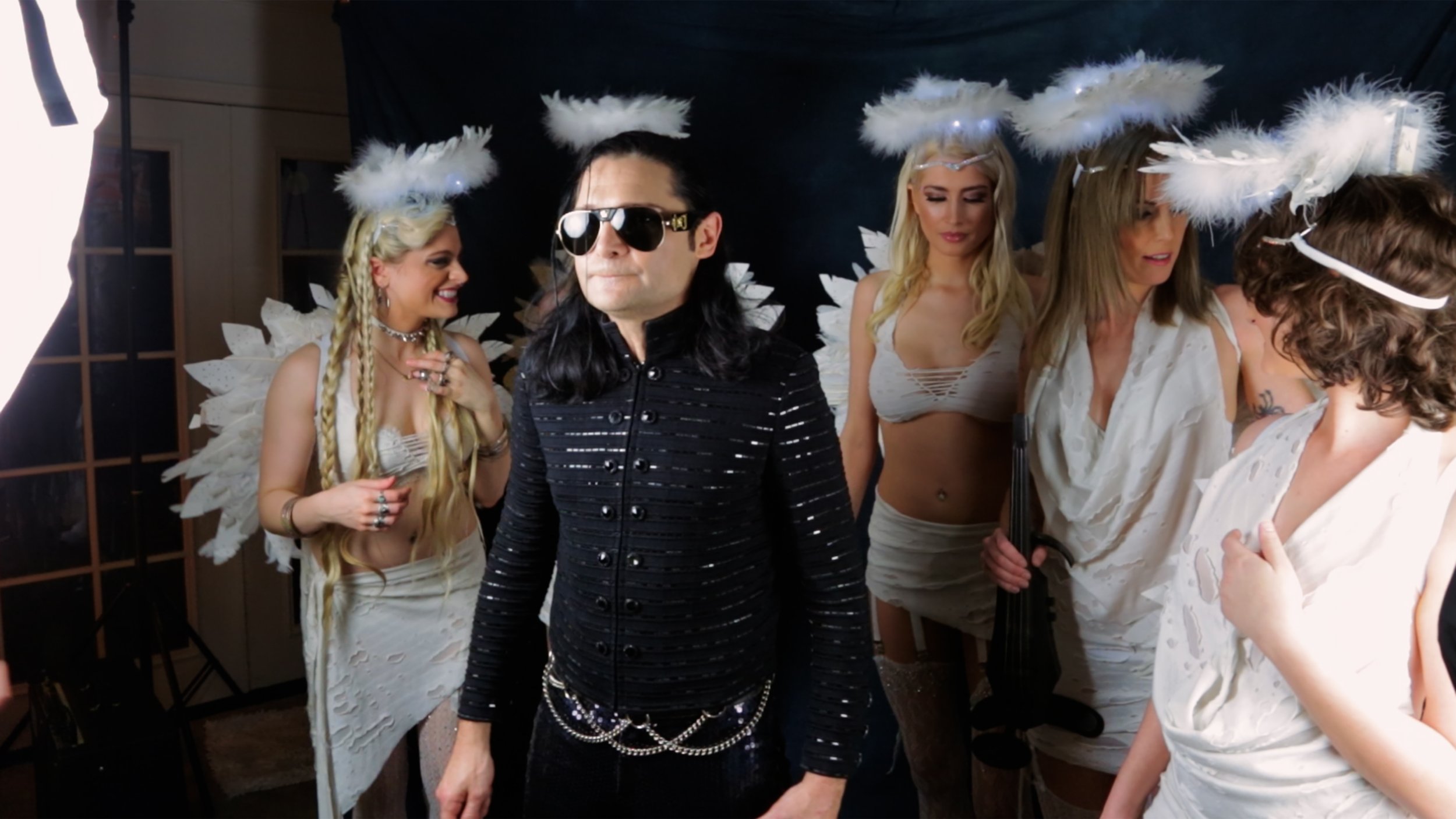 Corey Feldman, in sunglasses and a black leather jacket, surrounded by four women dressed as angels with feathered wings and halos, in a casual indoor studio setting.