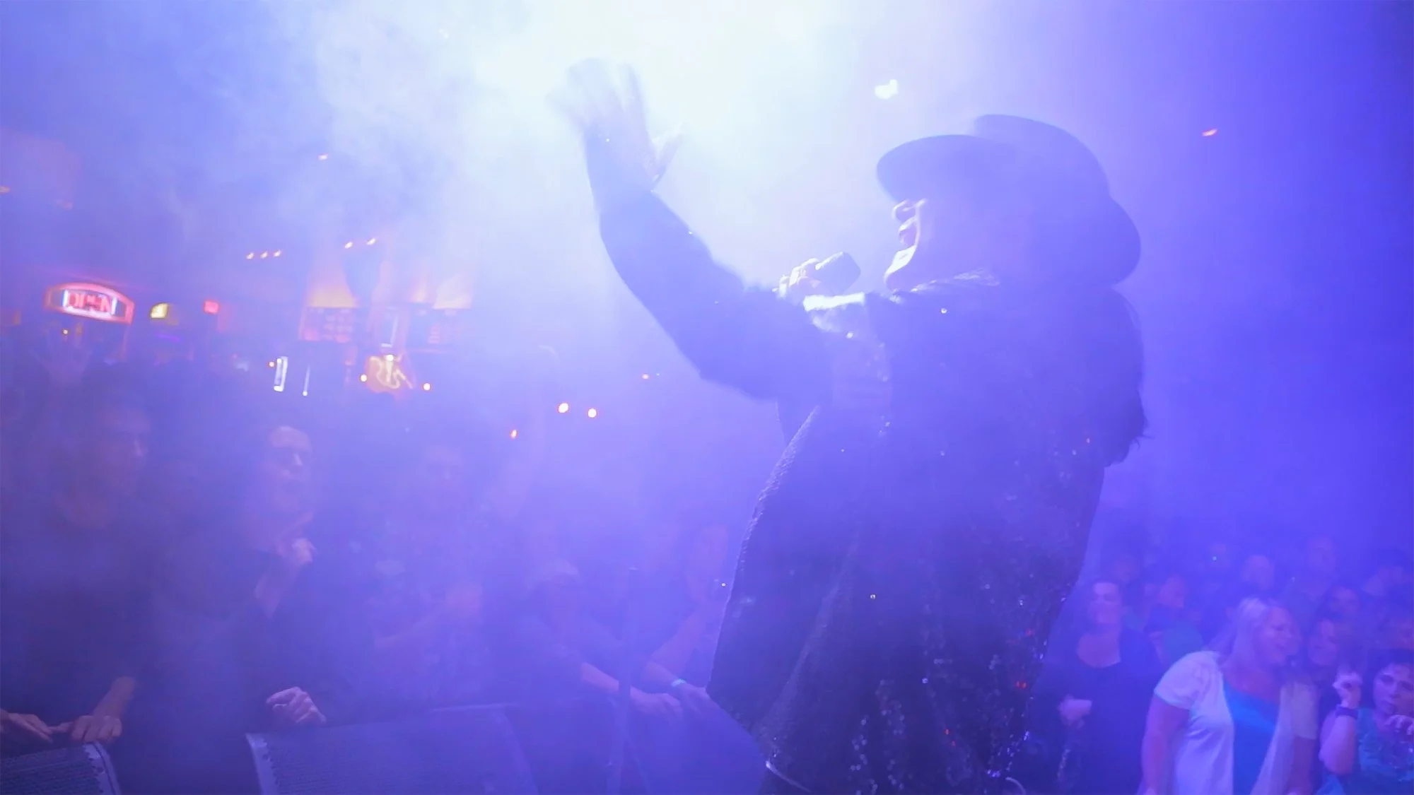 Corey Feldman singing into microphone on stage with bright light shining down, audience watching in a dark club
