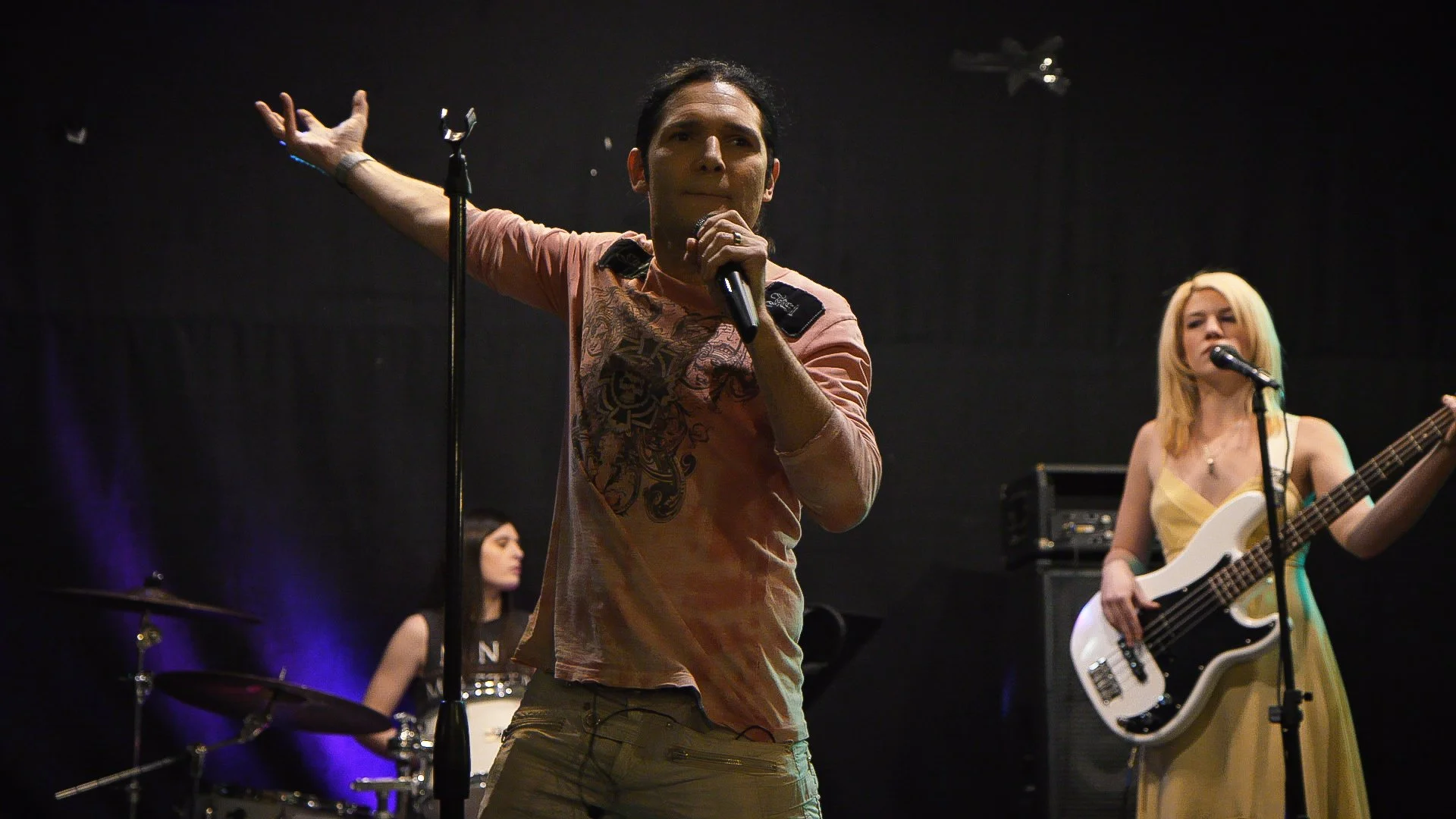 Corey Feldman performs on stage, with a male vocalist in the foreground, a female bassist on the right, and a female drummer in the background.