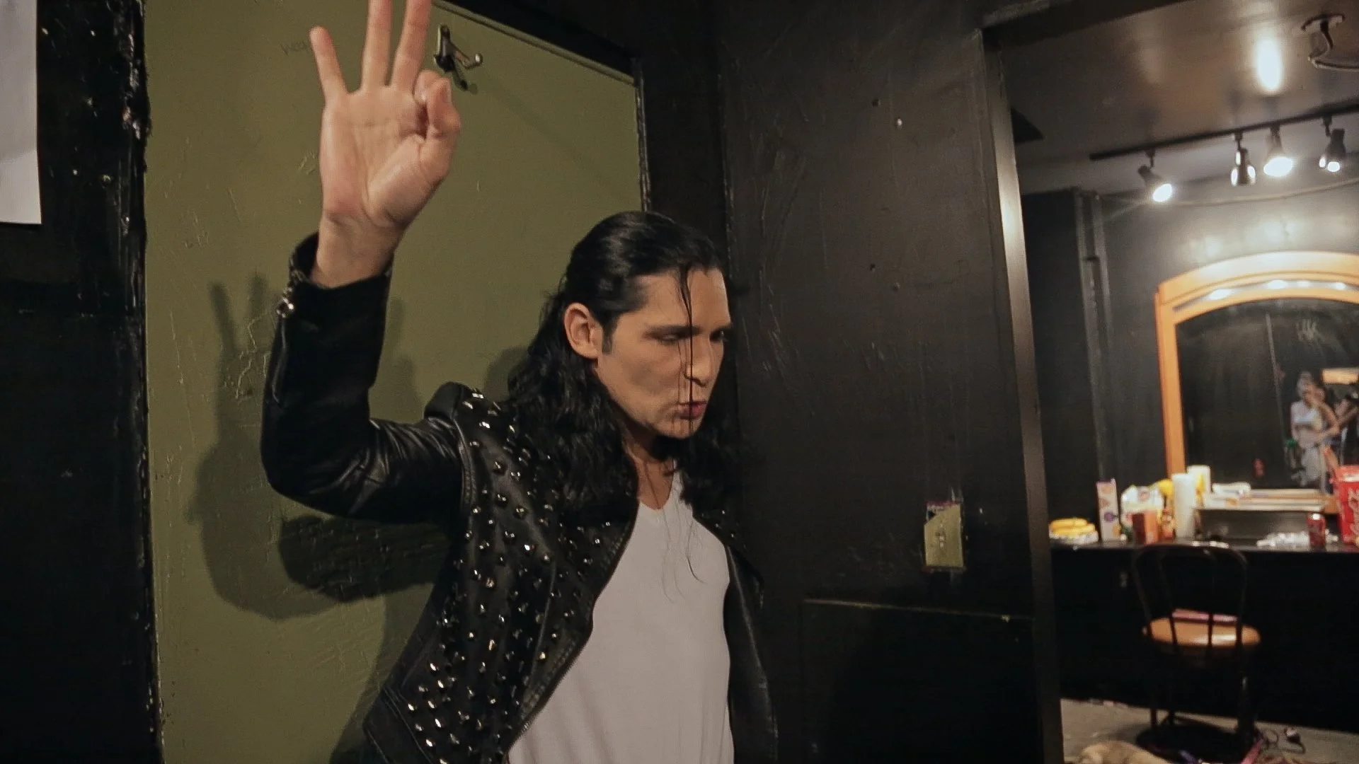 A person with long dark hair wearing a black leather jacket with studs stands against a green wall, with eyes closed and hand raised in a gesture, in a dimly lit room with a mirror and a cluttered table.