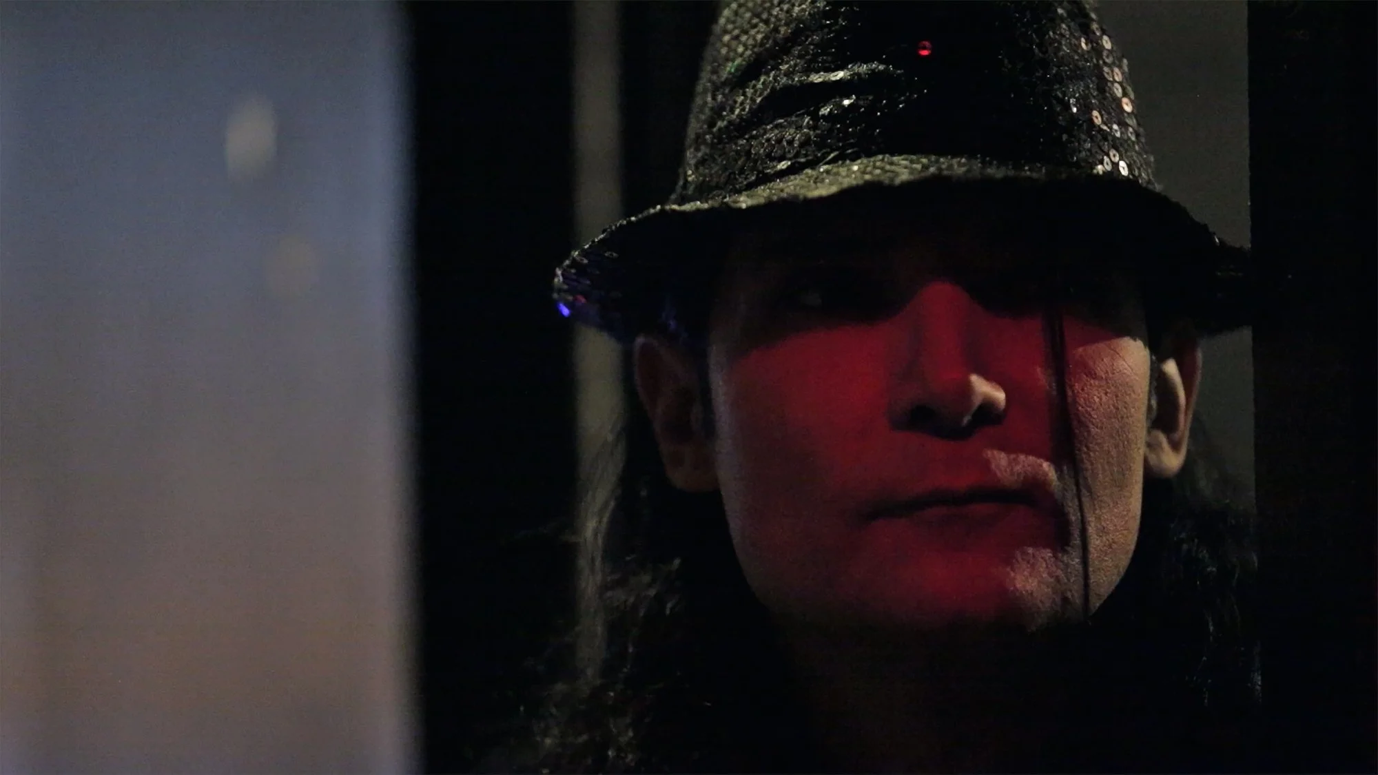 Corey Feldman wearing a shiny, sequined hat and dark glasses, with a dark background and red lighting on their face.