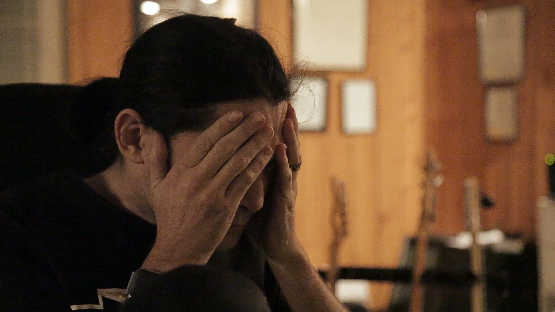 Corey Feldman sits indoors with a wooden wall and framed pictures in the background.