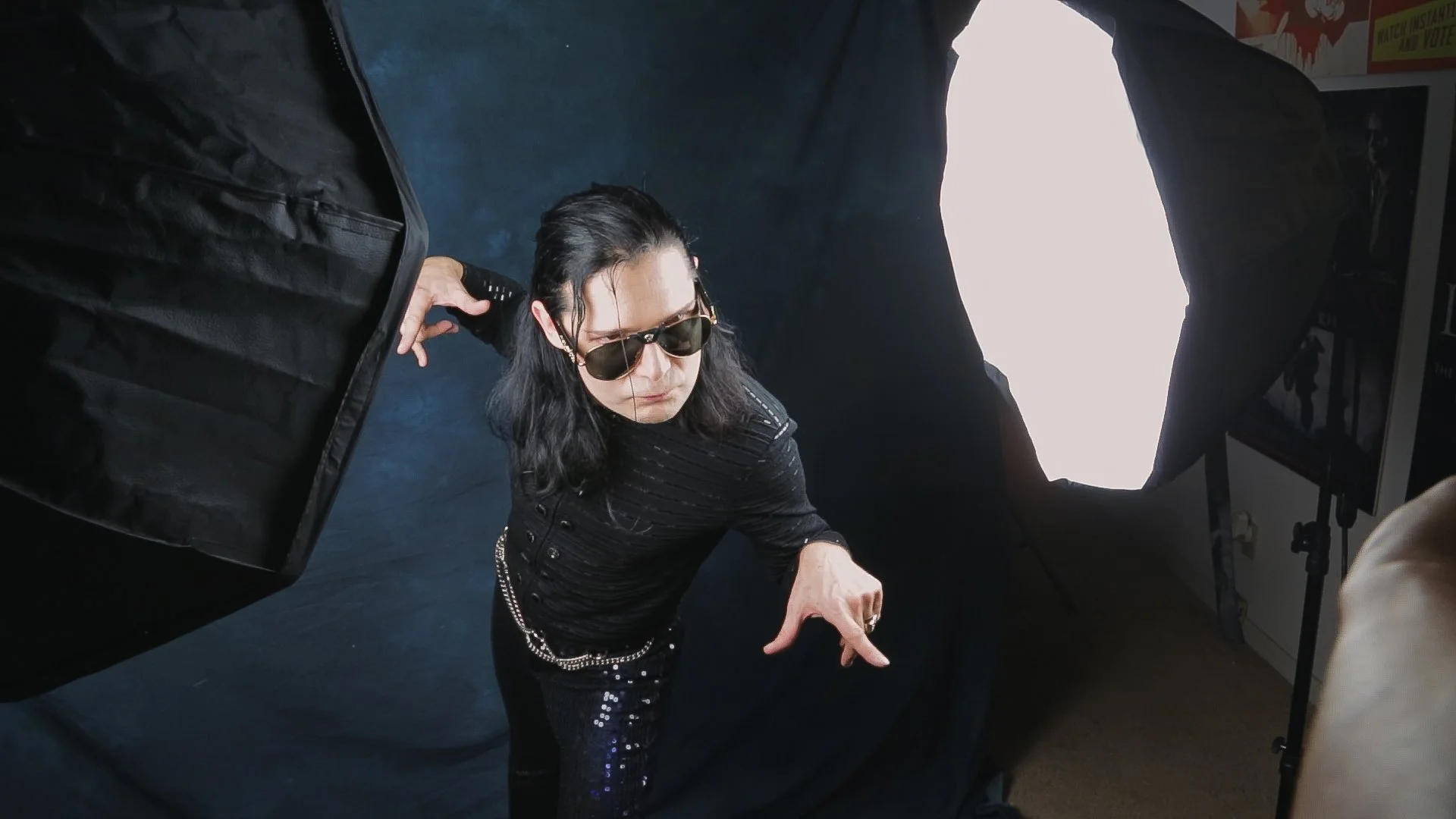 Corey Feldman wearing sunglasses and black clothing, is posing in a photo studio with a professional light setup.