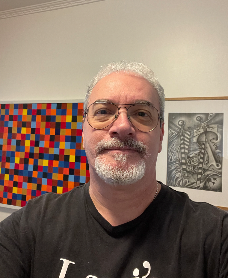 Homem de cabelo grisalho, barba por fazer, usando óculos com lente amarela, camiseta preta, tirando selfie diante de obras de arte na parede.