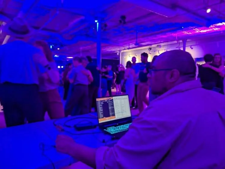 A photo looking over the shoulder of Omar as he sits at his laptop DJ'ing for an event. There is a blue light suffusing the scene and blurry dancers are visible in the background.