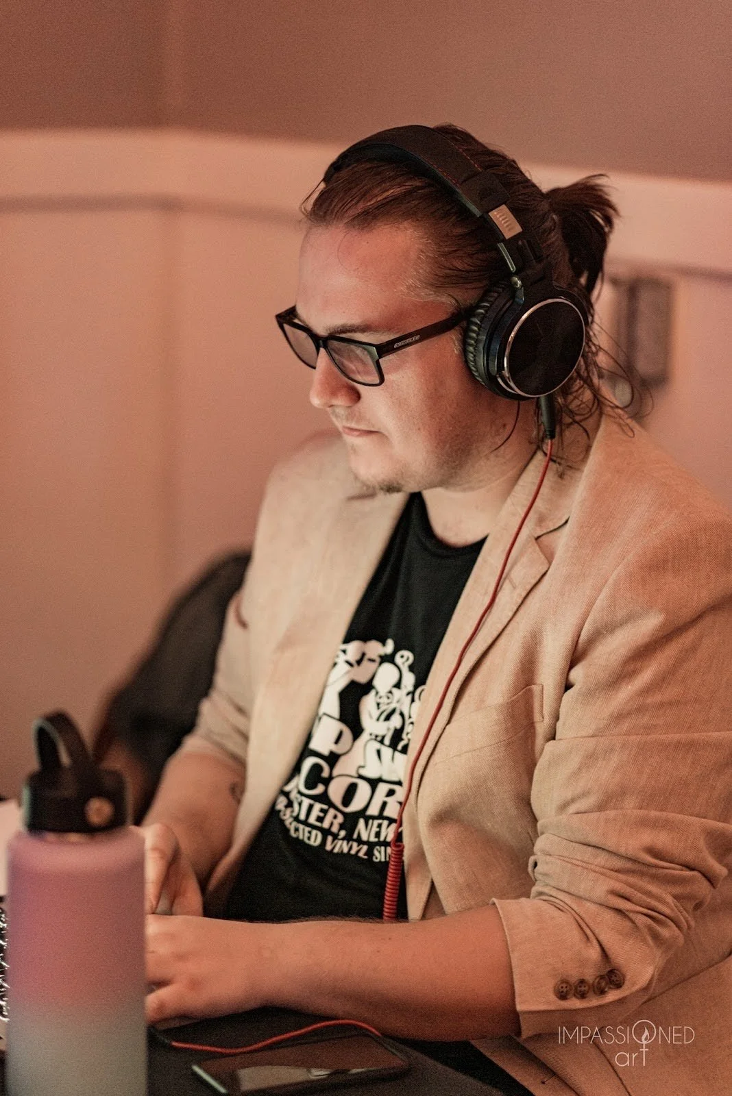 A photo of Art with headphones and a white suit jacket on. His shirt is from Bop Shop Records and he wears dark rimmed glasses, looking at his laptop.