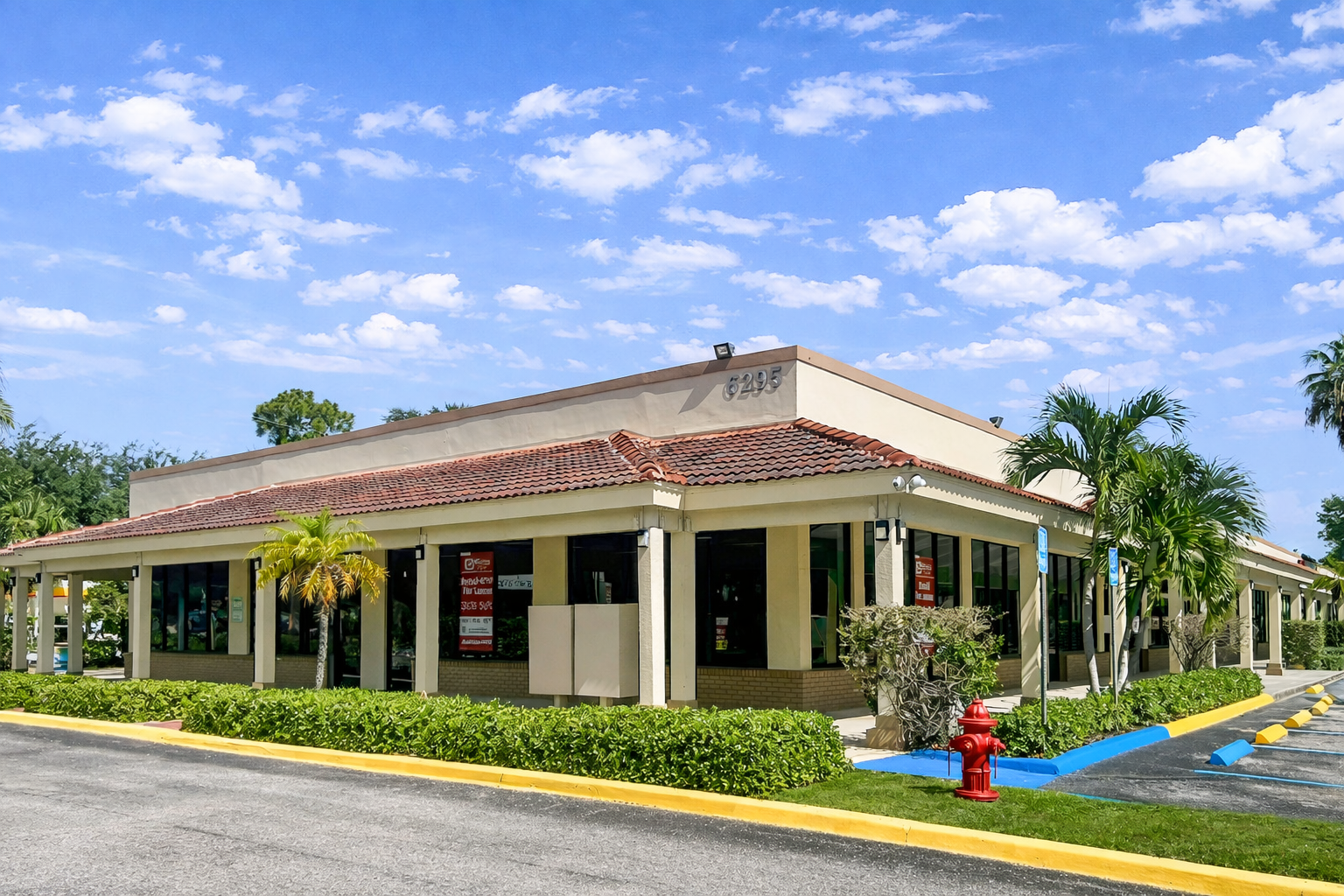 Exterior view of a commercial building with a red-tiled roof, beige walls, and large glass windows, surrounded by palm trees and greenery, with a parking lot in front.