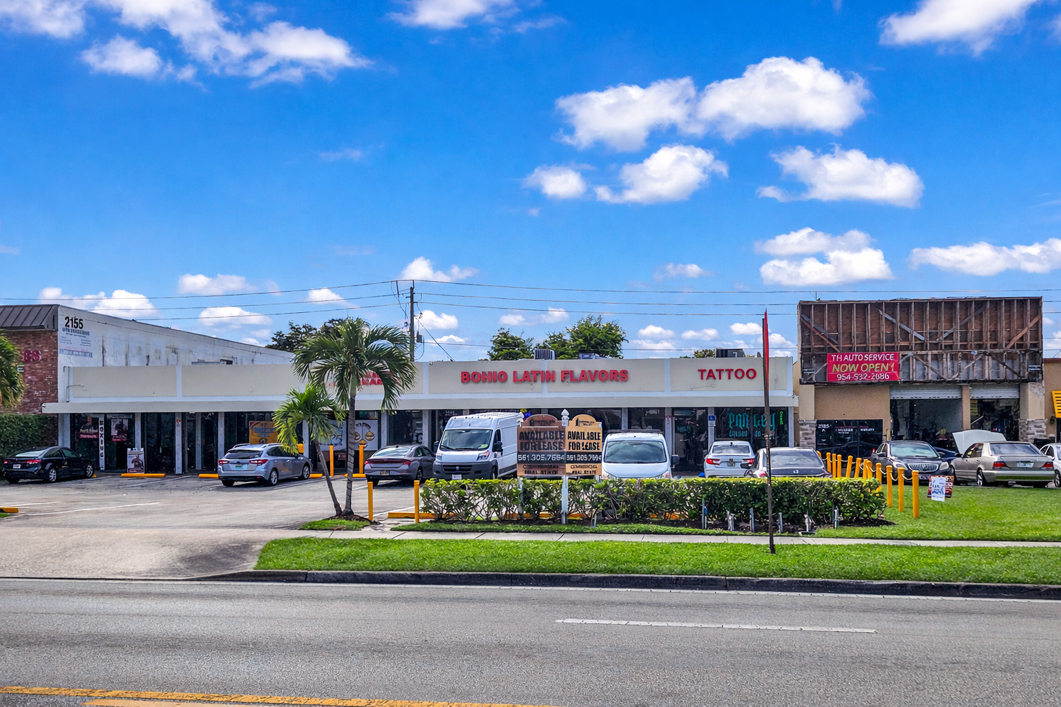 A strip mall with three storefronts and a parking lot in front. The signs read 'Bohio Latin Flavors,' 'Tattoo,' and a red banner advertising auto service. There are several cars parked and a small tree and bushes in the foreground.