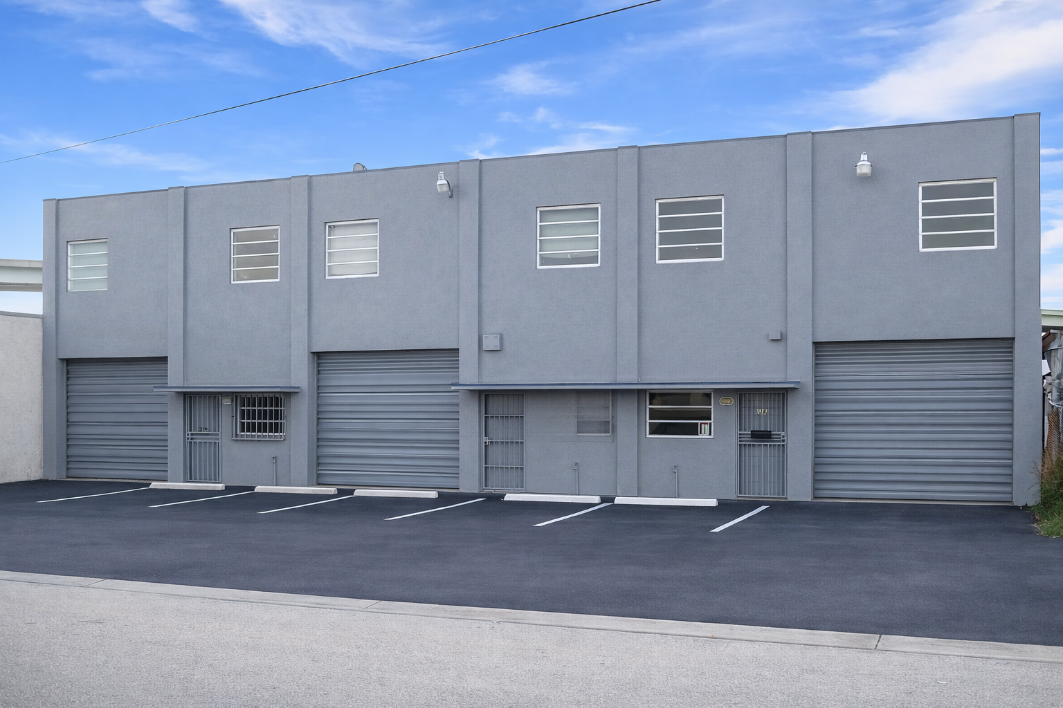 Storefront with multiple garage doors and small windows, painted gray, empty parking lot in front, clear blue sky.