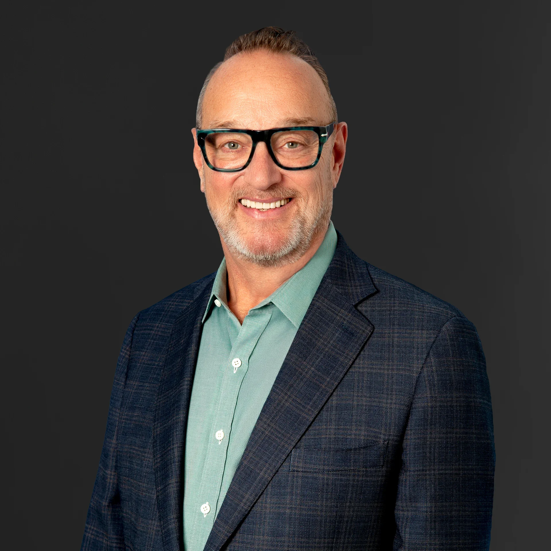 Headshot of a smiling middle-aged man with brown hair, glasses, and a beard, wearing a dark blazer and light green shirt against a dark background.