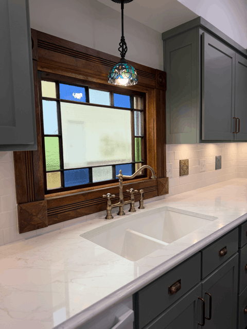 Victorian kitchen remodel sink area with white quartz countertops, brass faucet, and restored stained glass window with wood trim. 