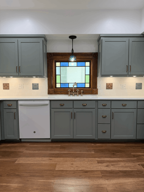  Kitchen sink wall with custom cabinetry, dishwasher, white tile backsplash, and restored stained glass window in historic remodel. 