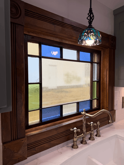  Close-up of restored stained glass kitchen window with brass faucet and quartz countertop in Victorian kitchen remodel. 
