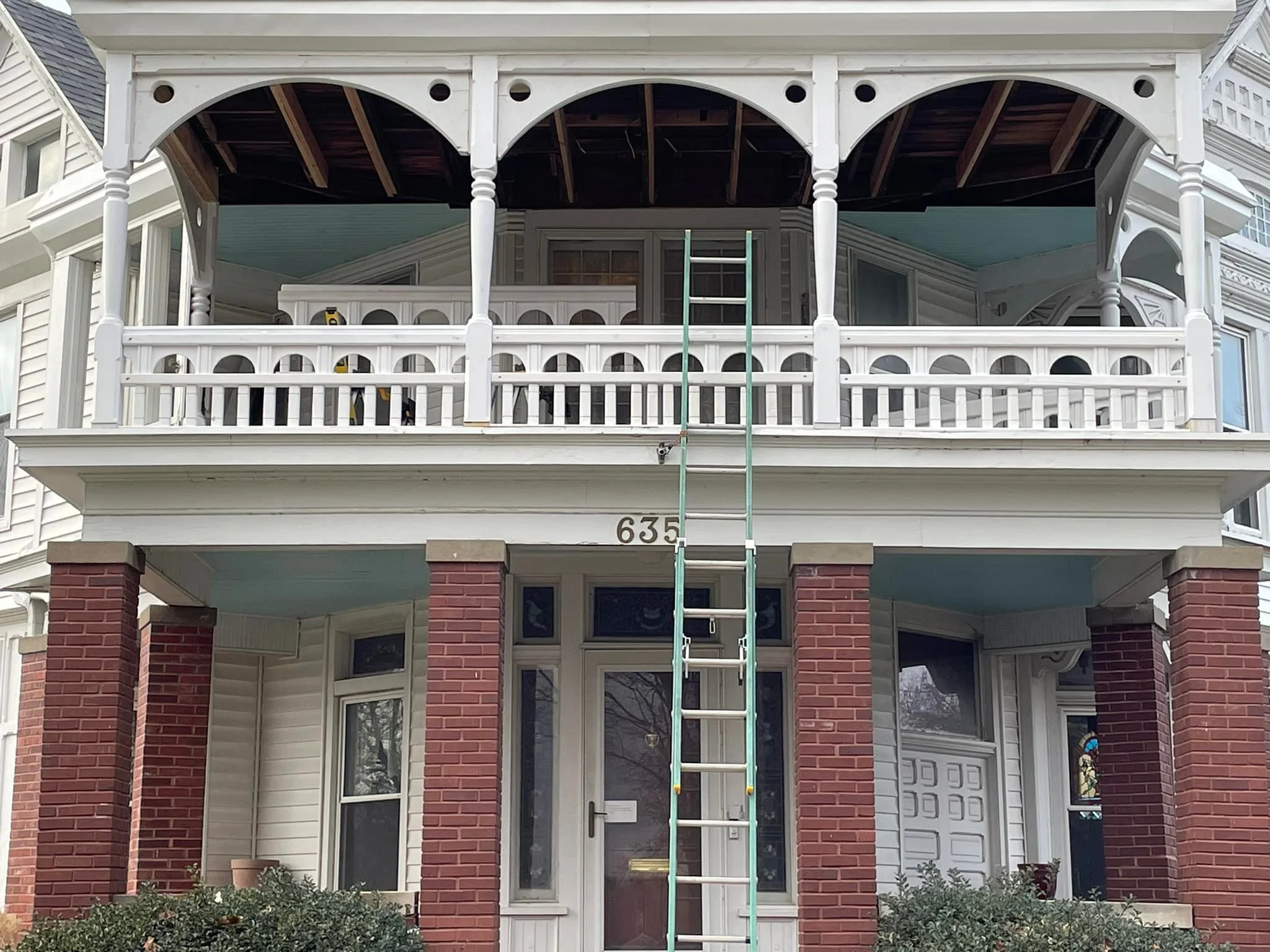 Front view of a building with a brick and siding facade, featuring a covered front porch and an upper balcony. A ladder is positioned in front of the house, reaching from the ground to the upper porch. The house number, 635, is visible above the entrance.