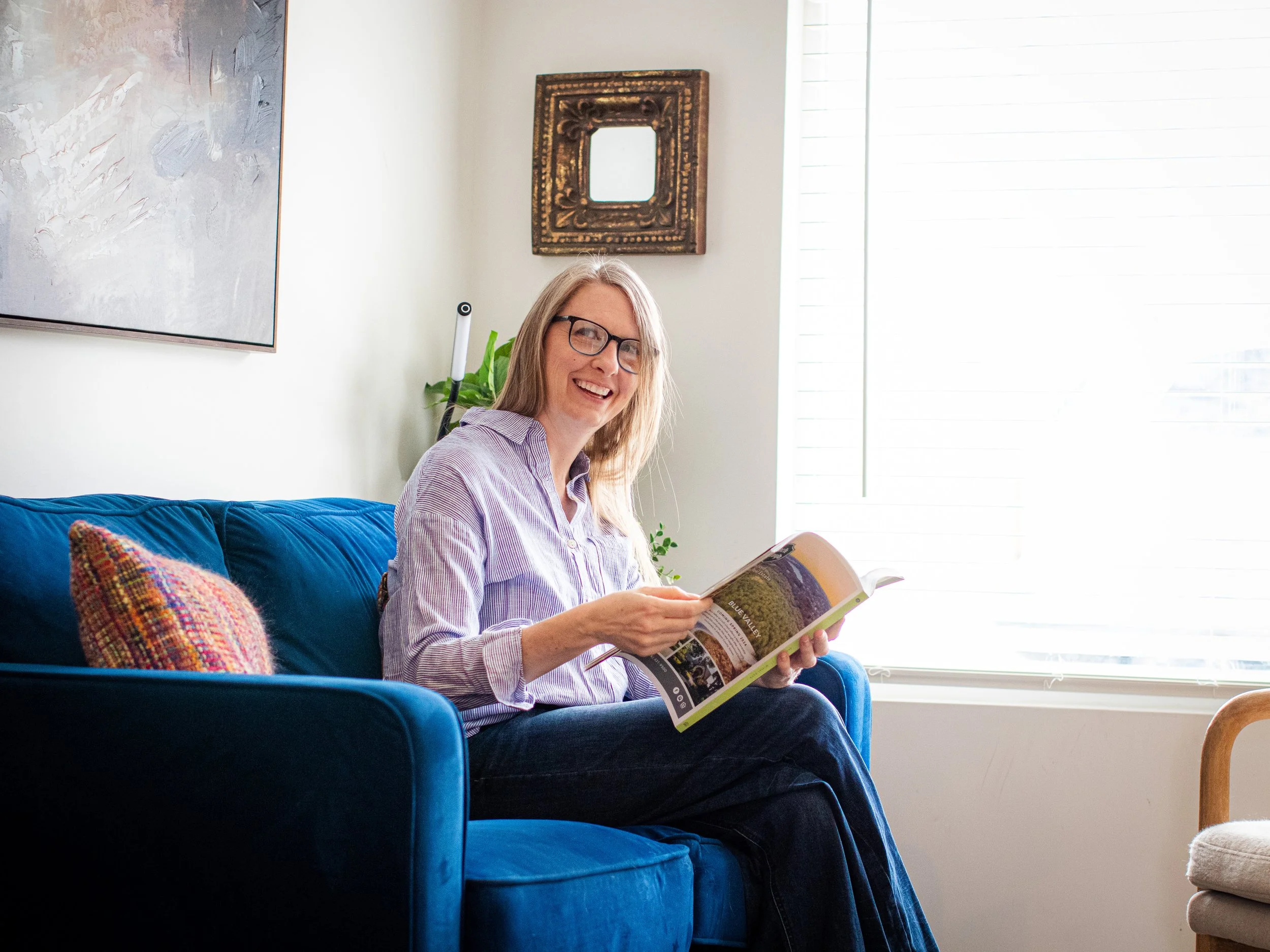 Amber Parker at a restyle appointment sitting in a blue velvet chair by a bright window, smiling while holding a home design magazine in a modern living room.