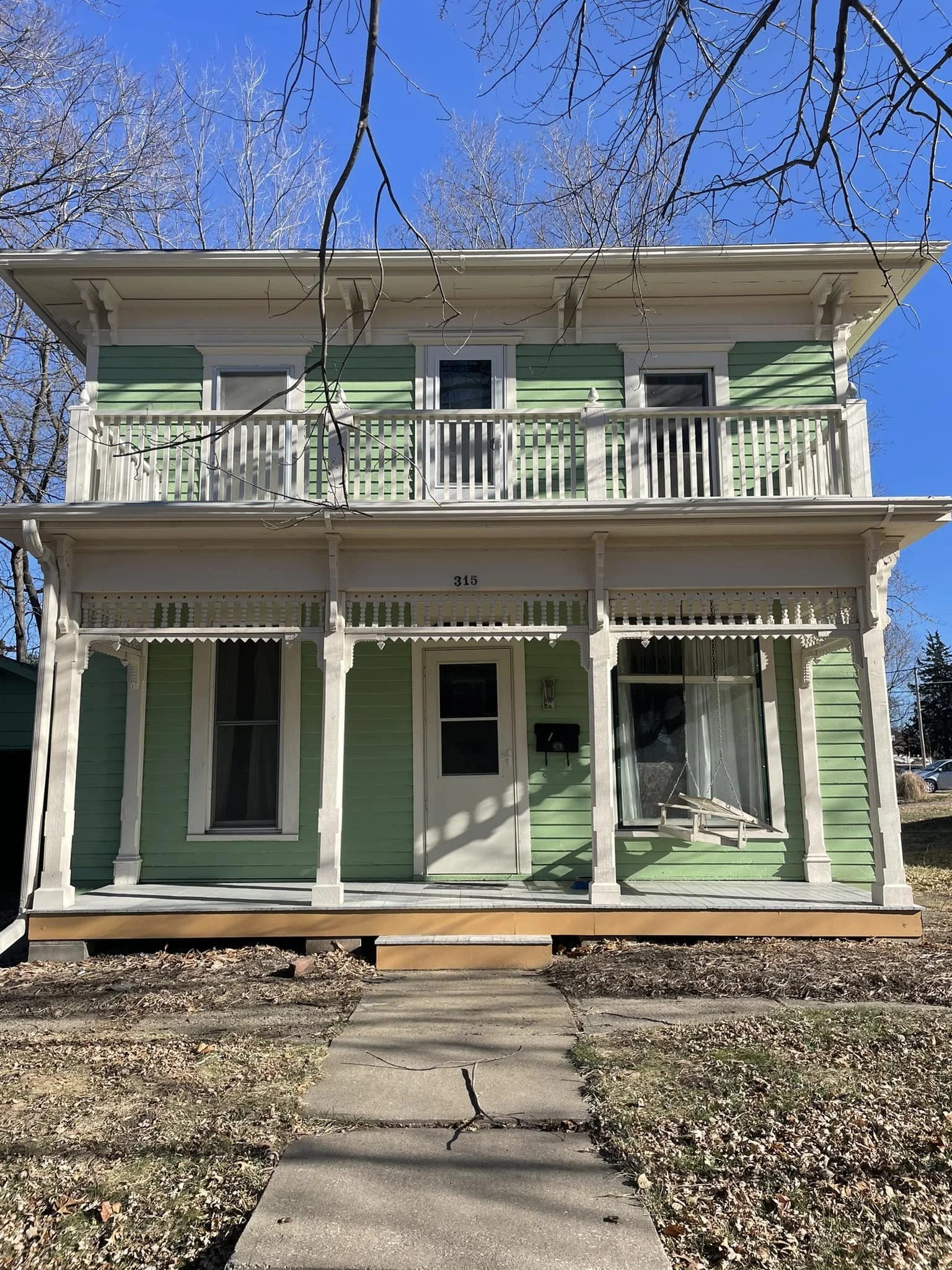 Before picture of a home renovation on a two-story green house with white trim and porch, a swing hanging from the front window, and a walkway leading to the entrance, under a clear blue sky in Nodaway County, Missouri.