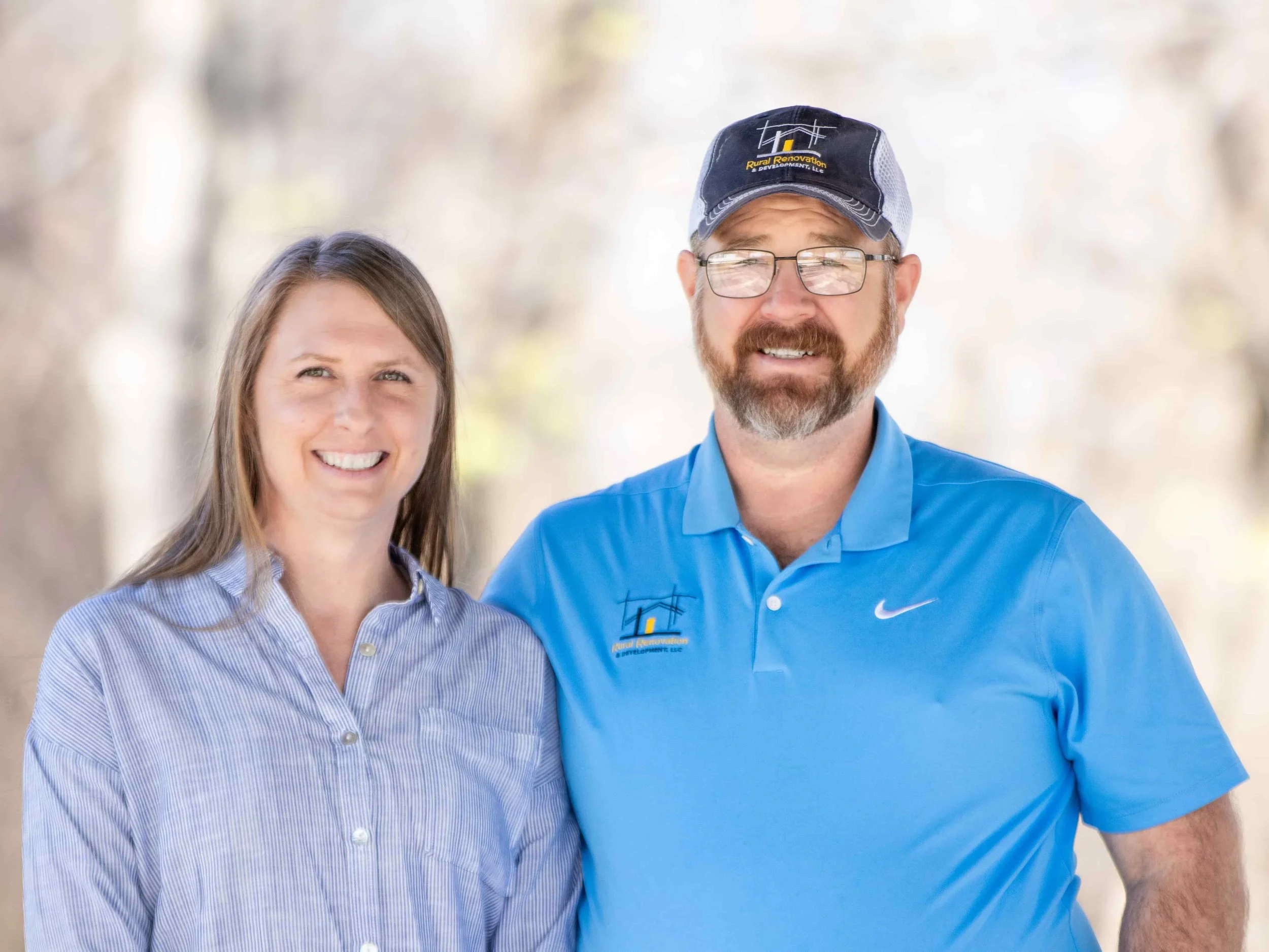 Amber and Matt smiling as they stand outdoors. Amber has long blonde hair and is wearing a blue and white striped button-up shirt. The man has a beard, glasses, and is wearing a blue polo shirt and a cap with a Rural Renovation shirt on.